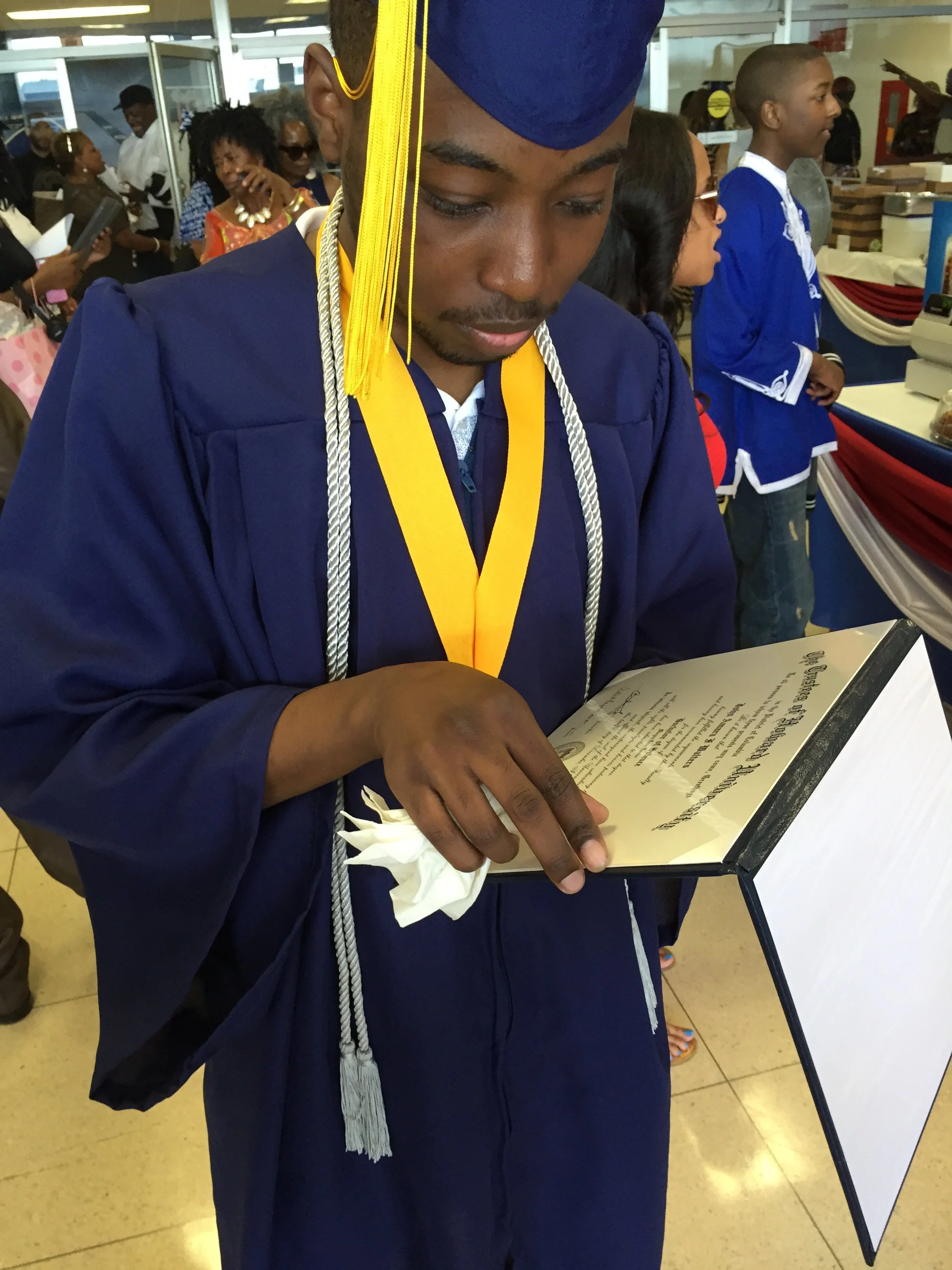 A young man in a navy blue graduation gown and cap with yellow cords and a white and gray honor cord, holding and reading a diploma or certificate, at a graduation ceremony with other graduates and family members in the background.