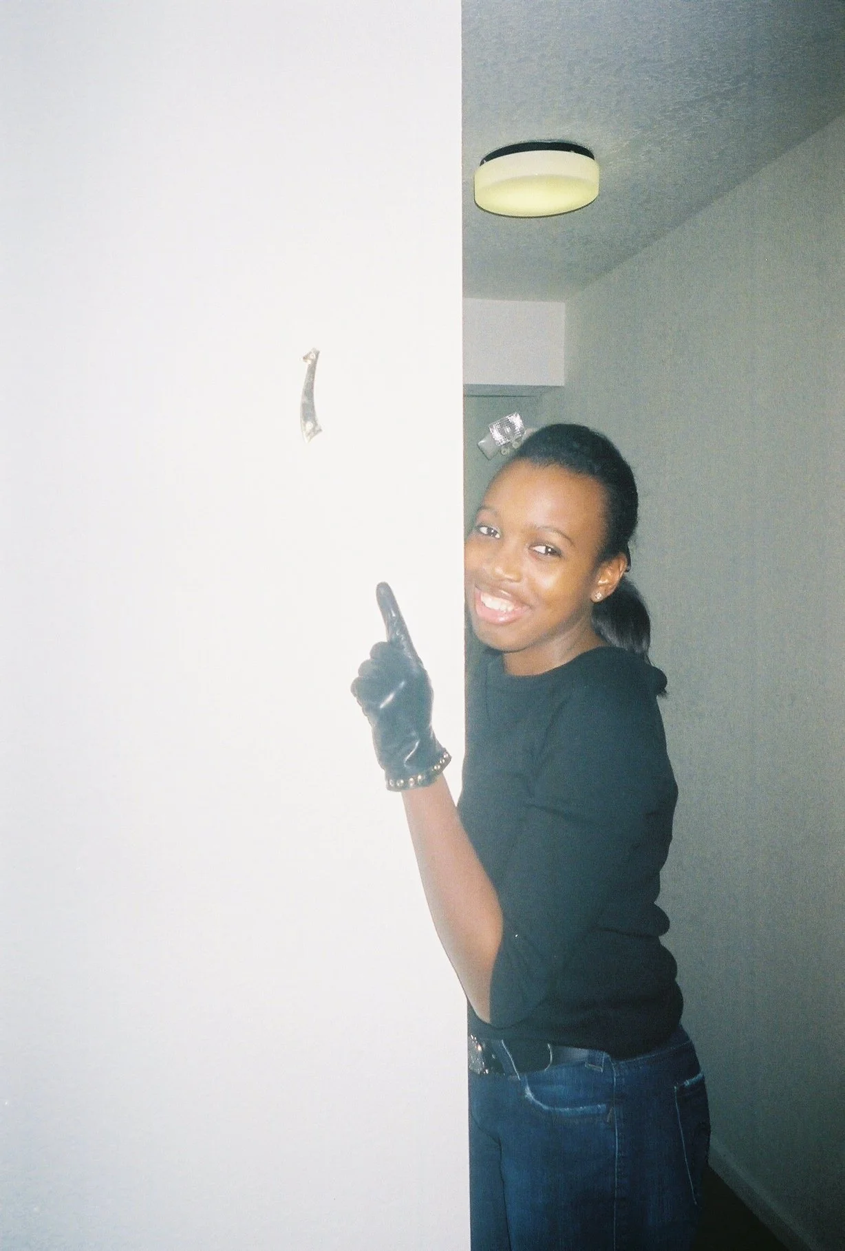 A young woman with a ponytail, wearing a black top, jeans, and a studded bracelet, peeking from behind a white wall, smiling and pointing upward.