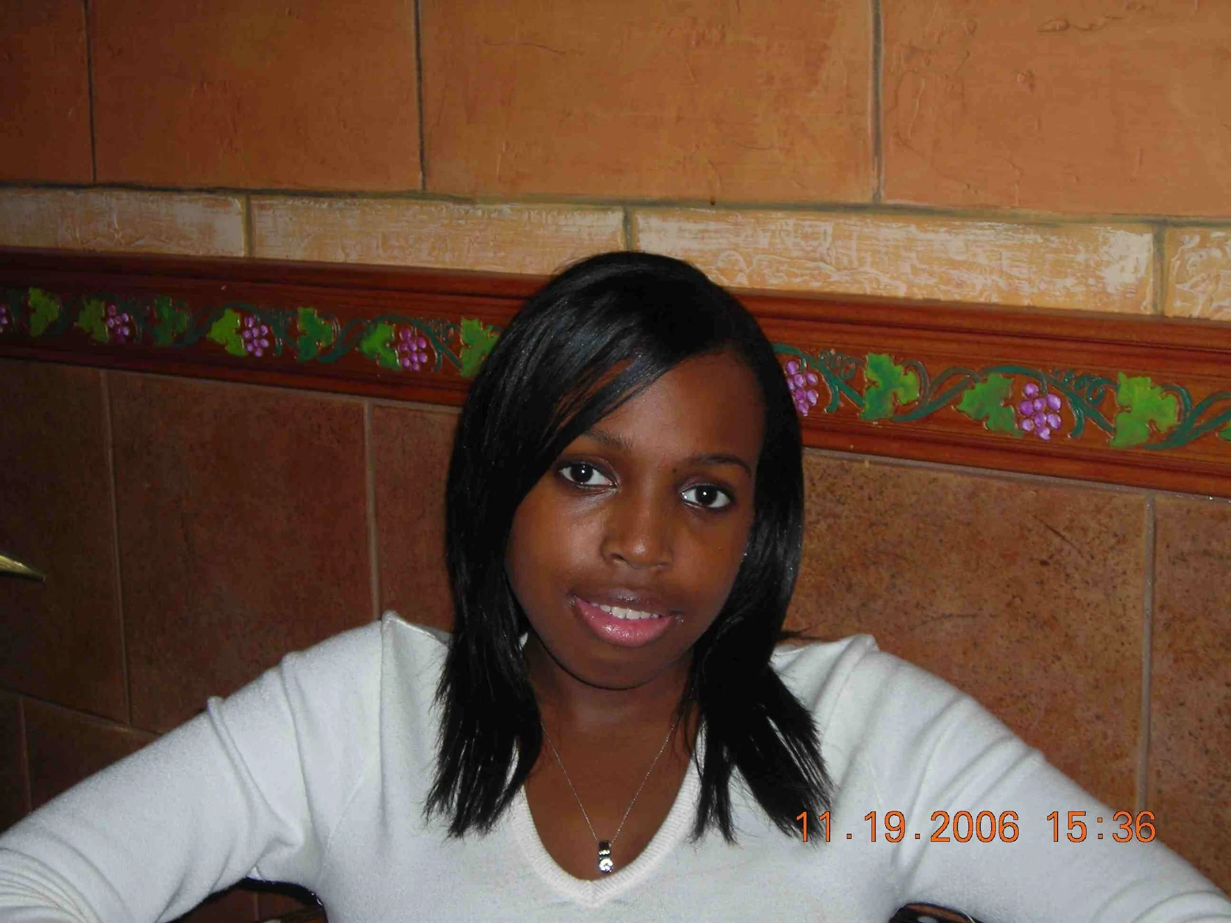 A young woman with straight black hair, wearing a white top and a necklace, smiling at the camera in an indoor setting with brown tile walls and decorative green and purple grapevine pattern.