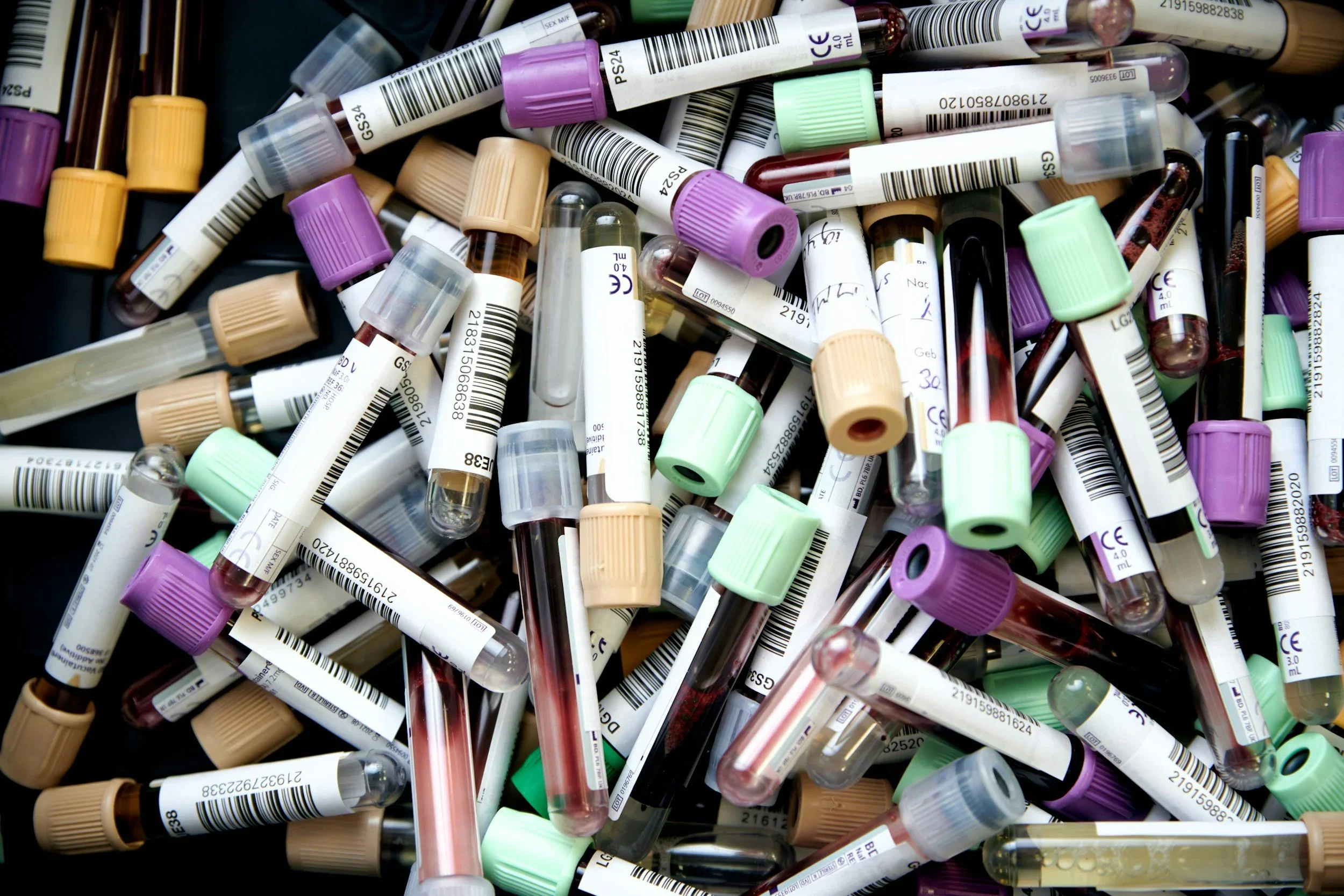 A large pile of used test tubes with various colored caps, including purple, green, and beige, some containing blood and others empty or with liquids.