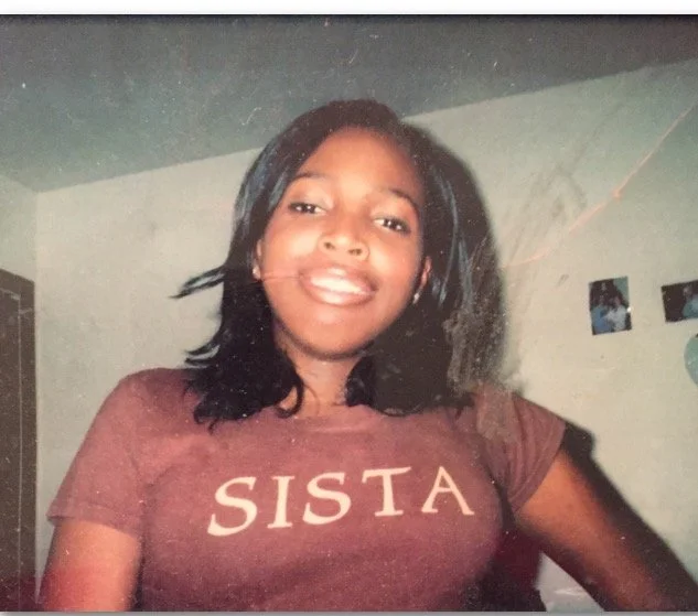 Young woman with shoulder-length black hair smiling, wearing a maroon T-shirt with the word 'SISTA' printed on it.