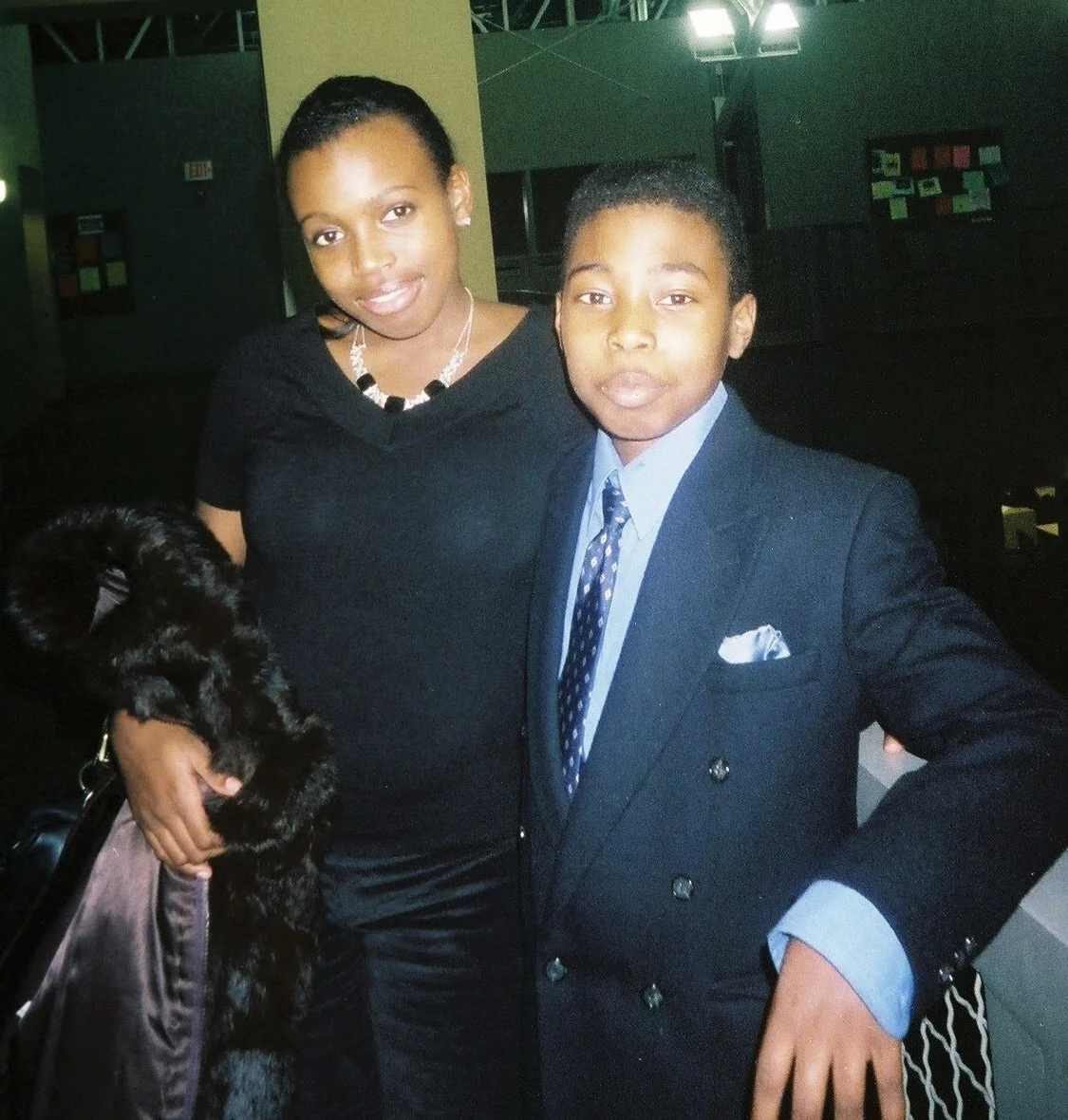 A young woman and young man posing together indoors. The woman is dressed in a black top with a necklace, holding a black bag with furry details. The man is wearing a dark suit with a blue shirt and a patterned tie, with a white pocket square. They are standing close together, smiling slightly, with a dark background and some lights visible.