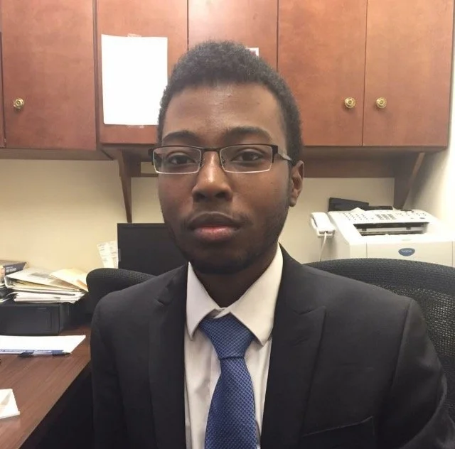 A young man in a suit and tie sitting in an office, with wooden cabinets, a printer, and papers in the background.