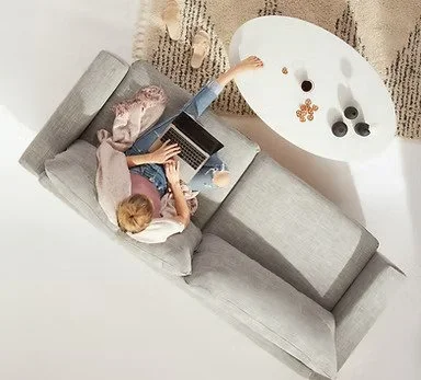 A woman lying on a gray sofa, holding a tablet, with a dog nearby, next to a white circular table with scattered coins and bottle caps.