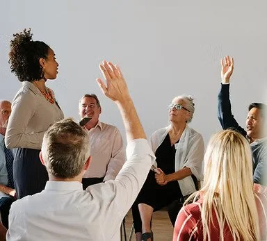 A diverse group of people in a meeting room, with some raising their hands and one person standing at the front speaking.
