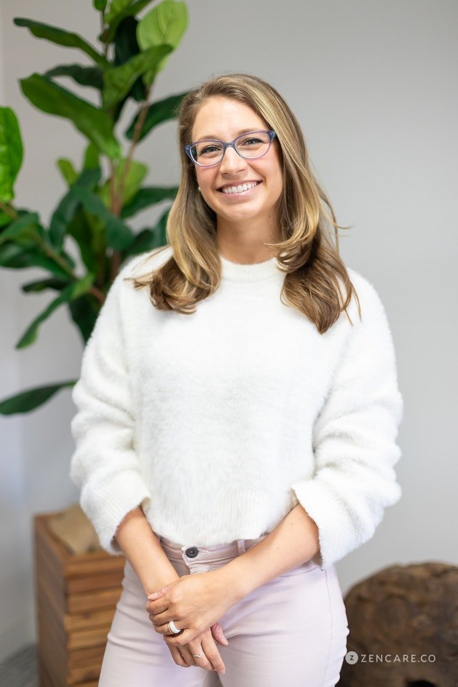 Dr. Patricia Pop, California psychiatrist offering telehealth in California. A smiling woman with glasses, wearing a white sweater and light-colored pants, standing in front of a large green plant in an indoor space.