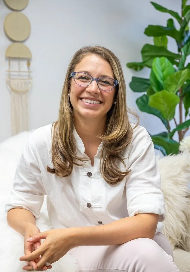 Holistic and Psychedelic Psychiatrist, Dr. Patricia Pop, wearing with glasses and long hair sitting on a white couch with a potted plant and wall hanging in the background.