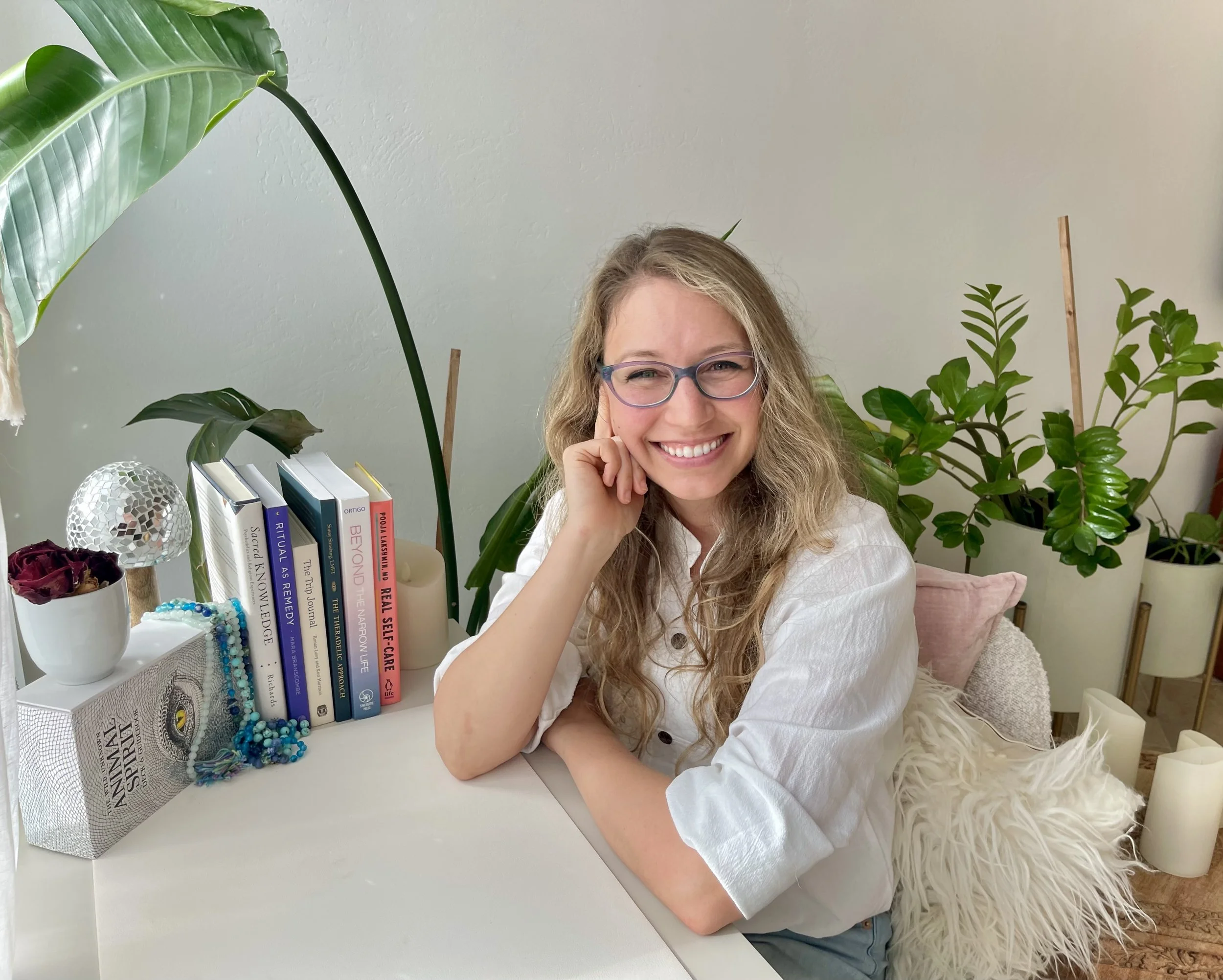 Dr. Patricia Pop, online psychiatrists in Palo Alto, Burlingame, and across California. A woman with long curly hair and glasses smiling while sitting at a white desk with books, decorative items, and a large leafy plant in the background.
