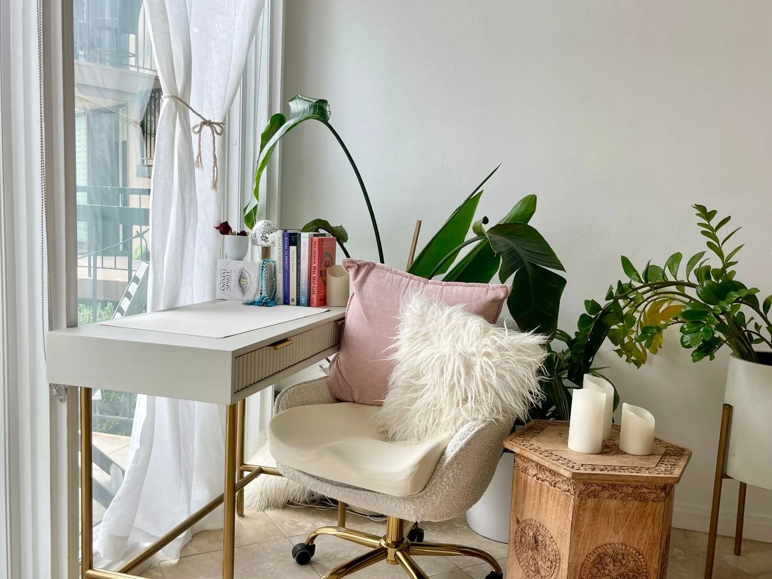 Psychiatrists Desks - A cozy corner of a room with a white desk near a window, decorated with books, a jewelry box, and a candle. An elegant chair with pink and white pillows is next to the desk, surrounded by large green plants.