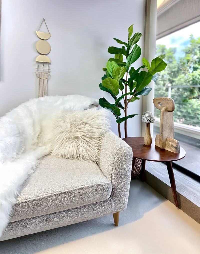 Comfortable therapist's office. Living room corner with a beige textured sofa, fuzzy white throw blanket, and matching pillow. Next to the sofa is a wooden side table with decorative wooden sculptures and a small mushroom-shaped lamp.