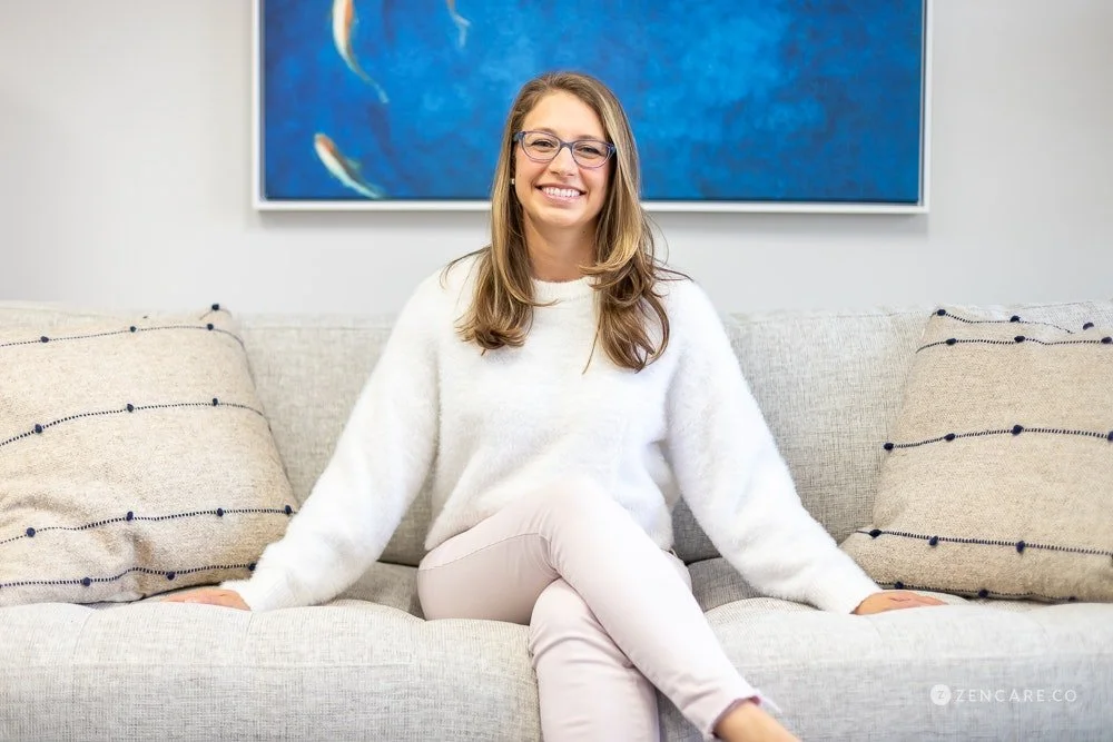 Dr. Patricia Pop, psychiatrist and therapist in Palo Alto and Burlingame, CA. Photo of her smiling with glasses and long hair sitting on a light-colored sofa in front of a blue wall with artwork, smiling.
