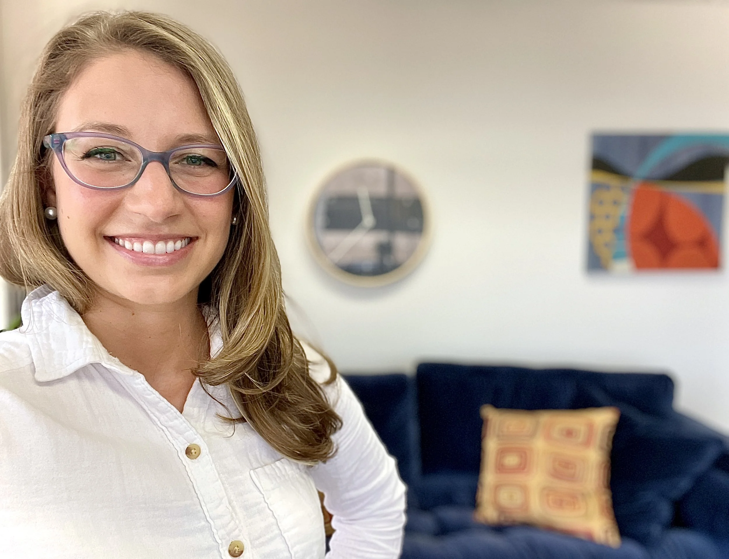 Dr. Patricia Pop, a holistic psychiatrist with blonde hair, glasses, and pearl earrings smiling in a white shirt, standing in a living room with a navy sofa and colorful artwork on the wall.