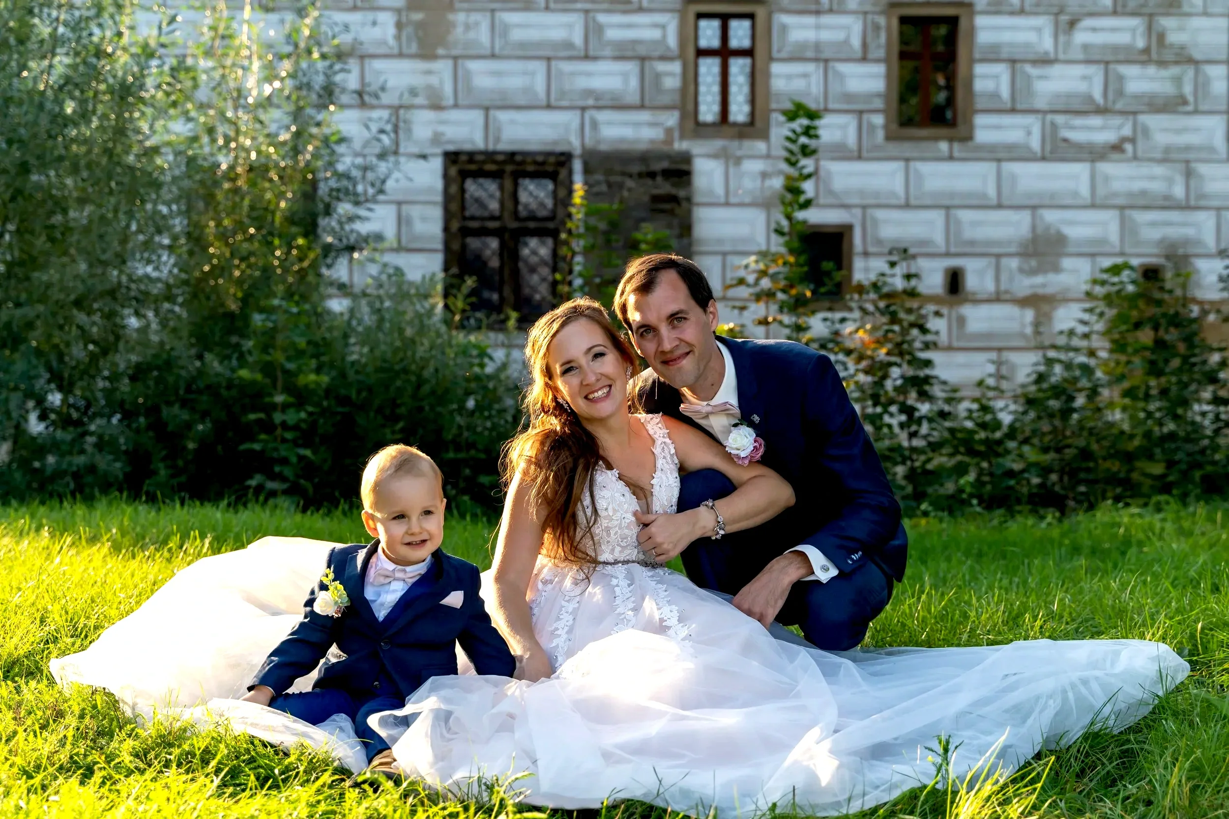 A newlywed couple in wedding attire, sitting outdoors on the grass with a young child, in front of a rustic building with small windows and green foliage.