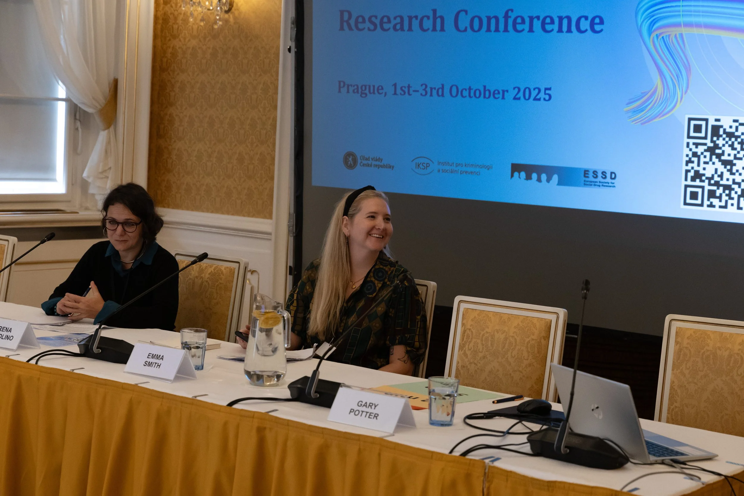 Two women seated at a conference table with microphones and name tags; a large screen behind them displays information about a research conference in Prague, October 2025.
