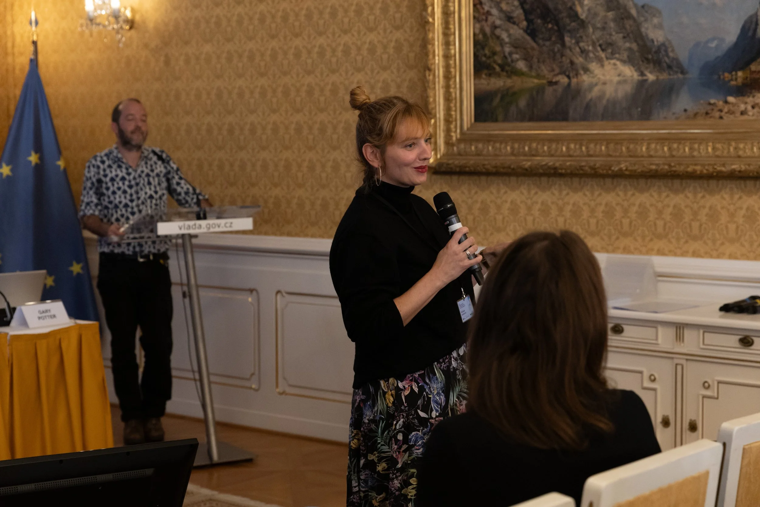 A woman holding a microphone speaks at a formal event in a room with ornate wallpaper, framed landscape painting, and an EU flag in the background.