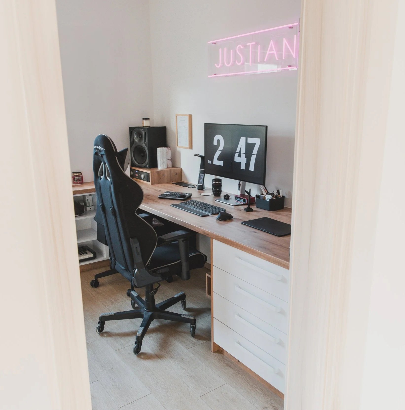A home office with a wooden desk, black ergonomic chair, computer monitor displaying the time 2:47 PM, speaker, office supplies, and a neon pink sign with the name 'Justian' on the wall.