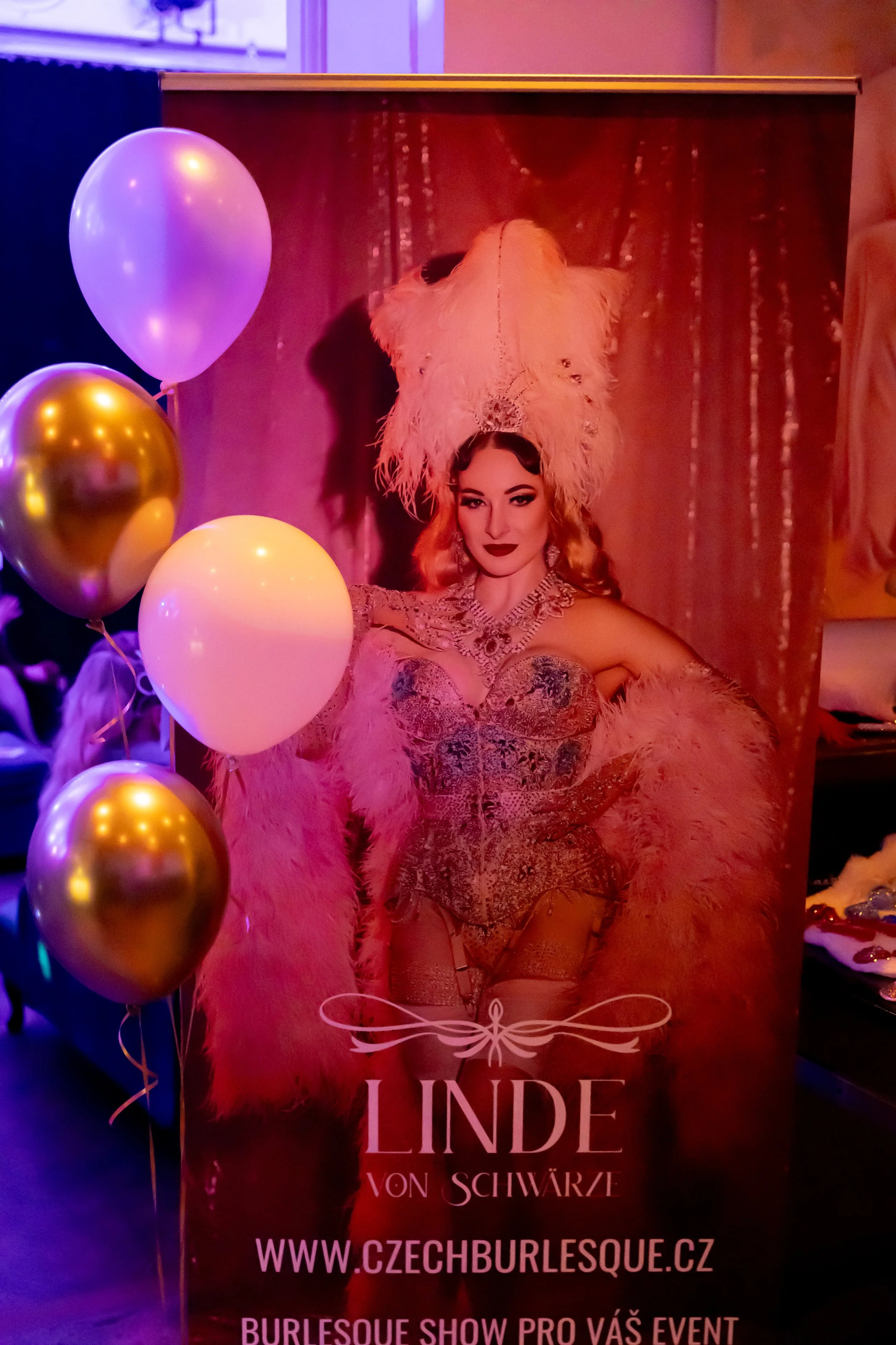 A poster of a burlesque performer dressed in elaborate costume with a feathered headpiece and jewelry, partially covered by balloons, promoting a show called "Linde von Schwarze" at czechburlesque.cz.