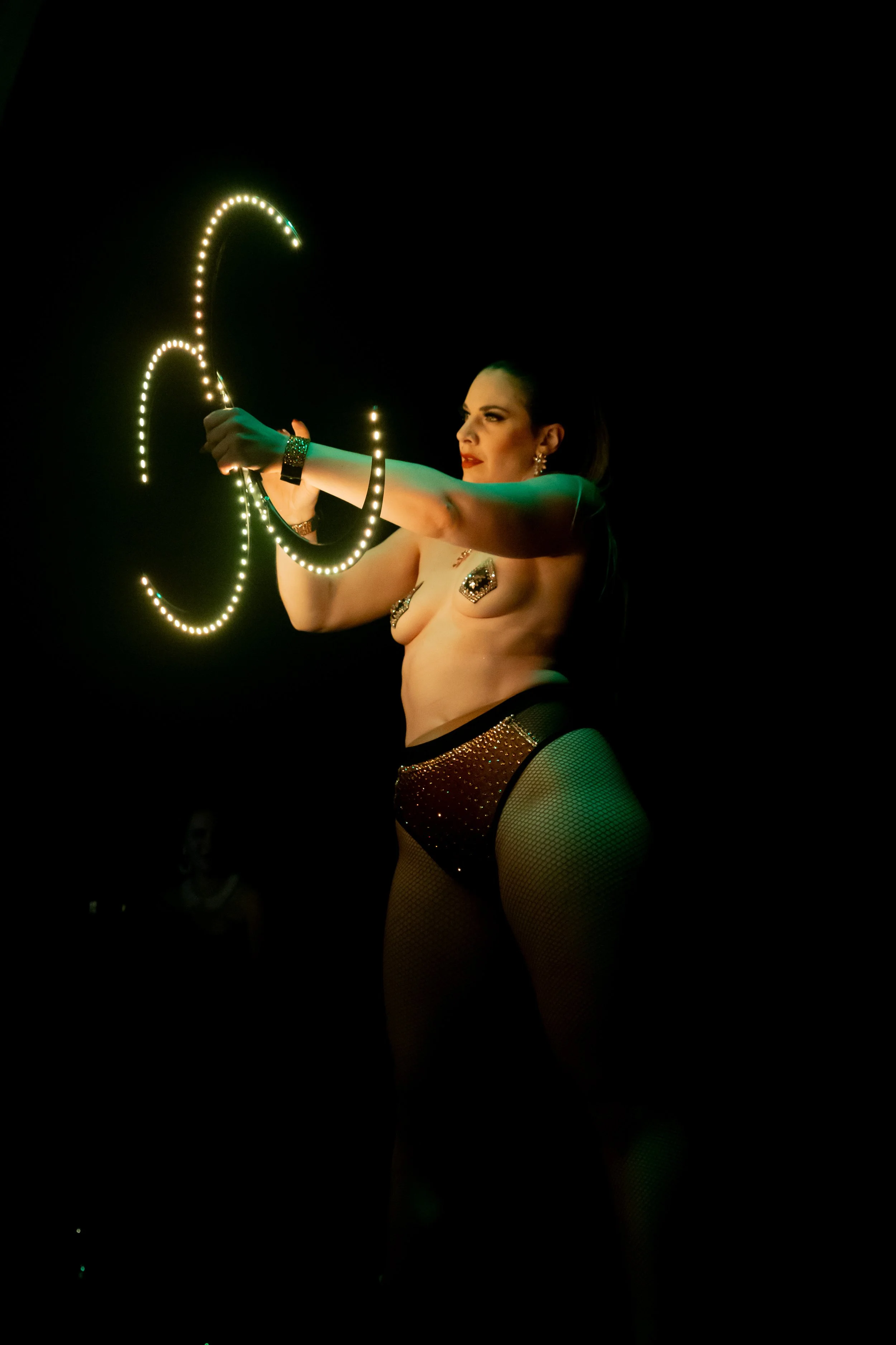 A woman performs a fire dance, spinning illuminated poi or staffs, with her body partially adorned with jewelry and fishnet stockings, against a dark background.