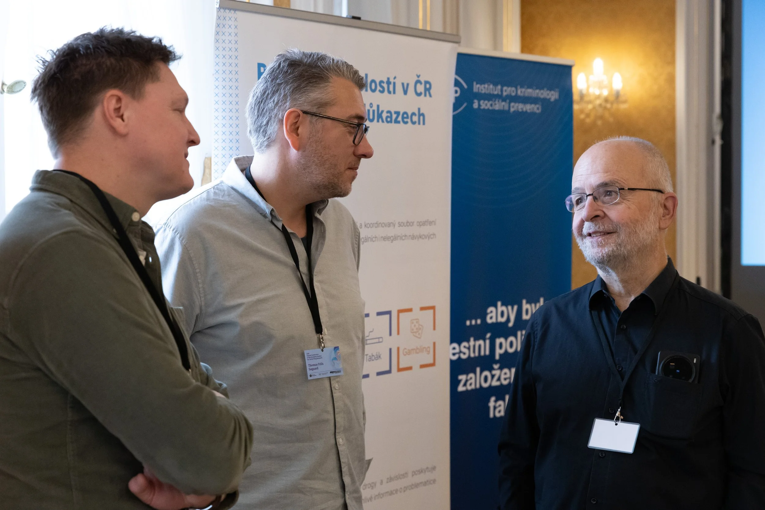 Three men in conversation at a conference, with banners and a chandelier in the background.