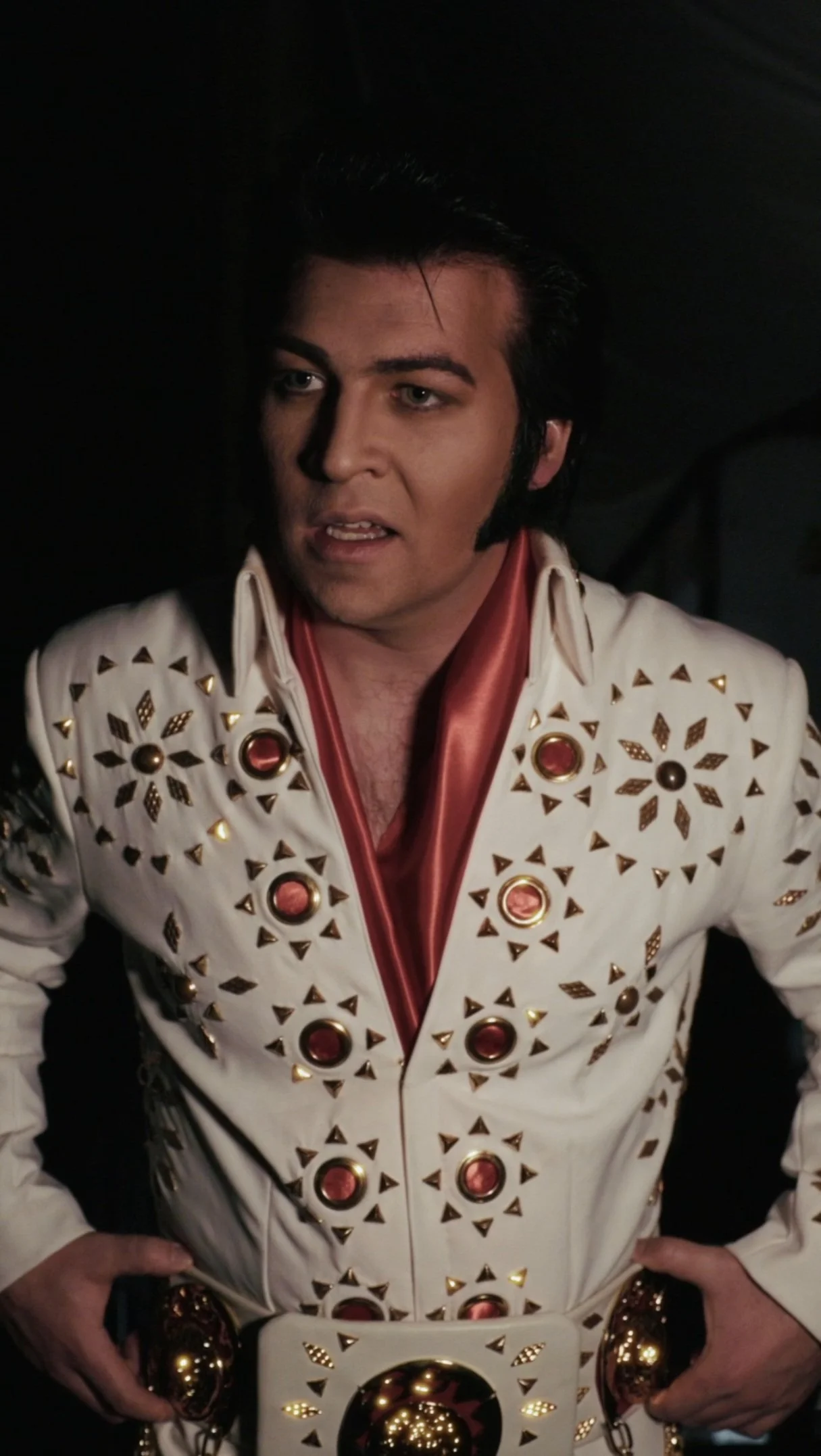 A man dressed as Elvis Presley wearing a jeweled white jumpsuit with red and gold accents.