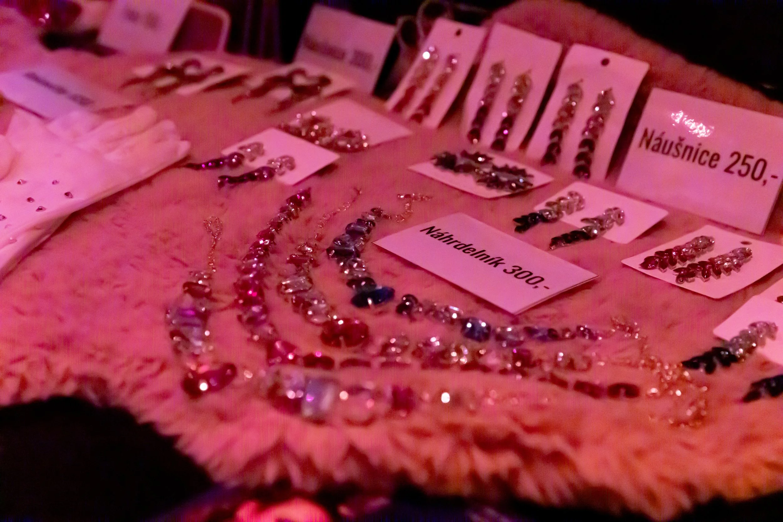 Display of jewelry on a pink, fluffy surface, including necklaces, earrings, and rings, with price tags labeled in Czech koruna.