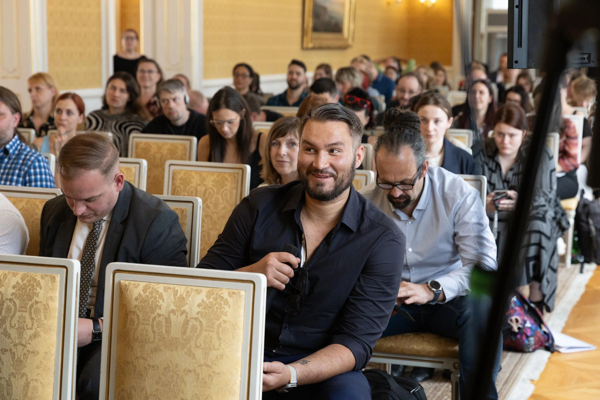 A man with a beard and dark shirt holding a microphone, smiling, seated in a large conference room filled with people attending a presentation or seminar.