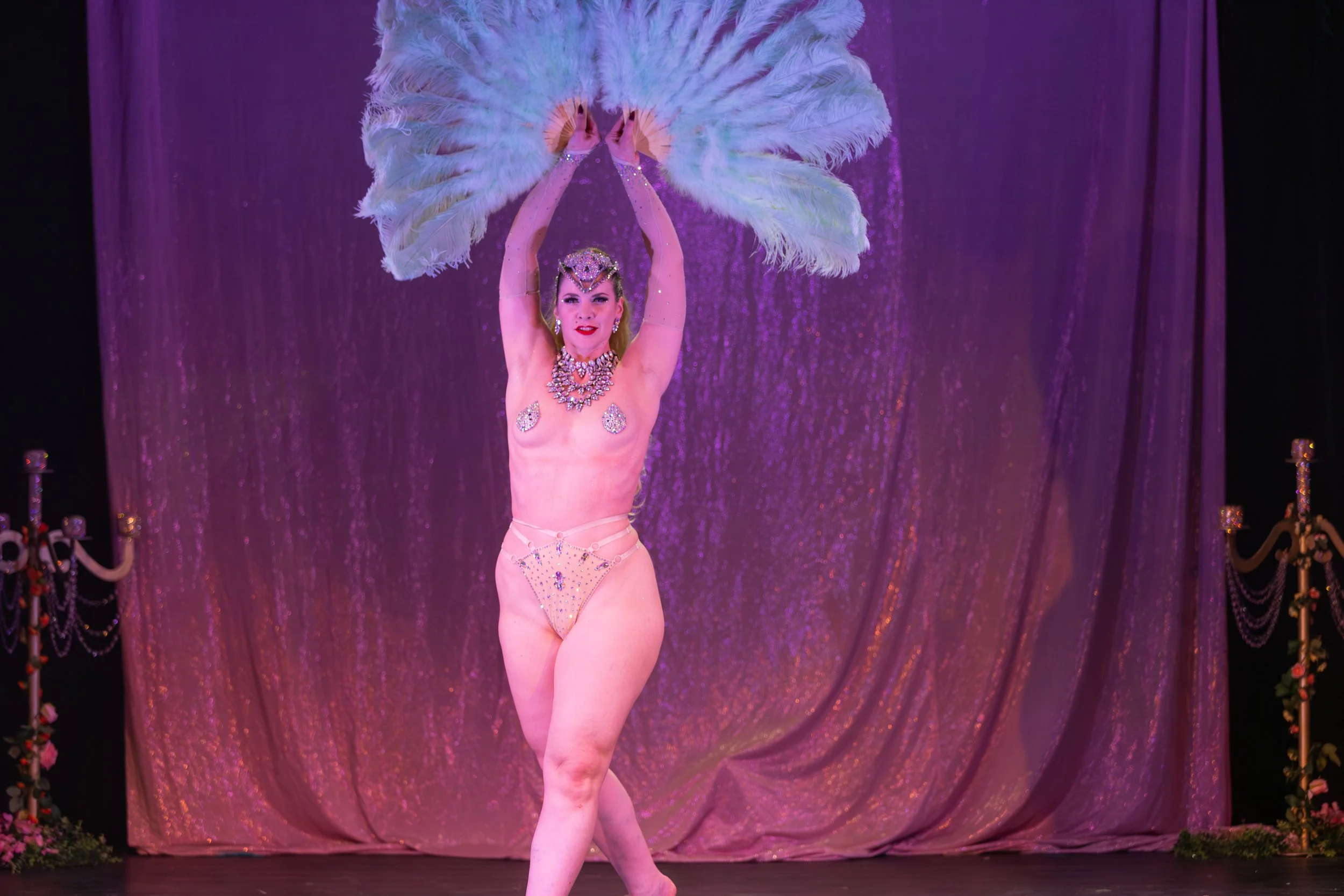A performer in a burlesque or cabaret costume on stage, holding large white feather fans above her head. She wears a jeweled headpiece, necklace, earrings, and a sparkling bikini, with a pink backdrop and stage decorations.