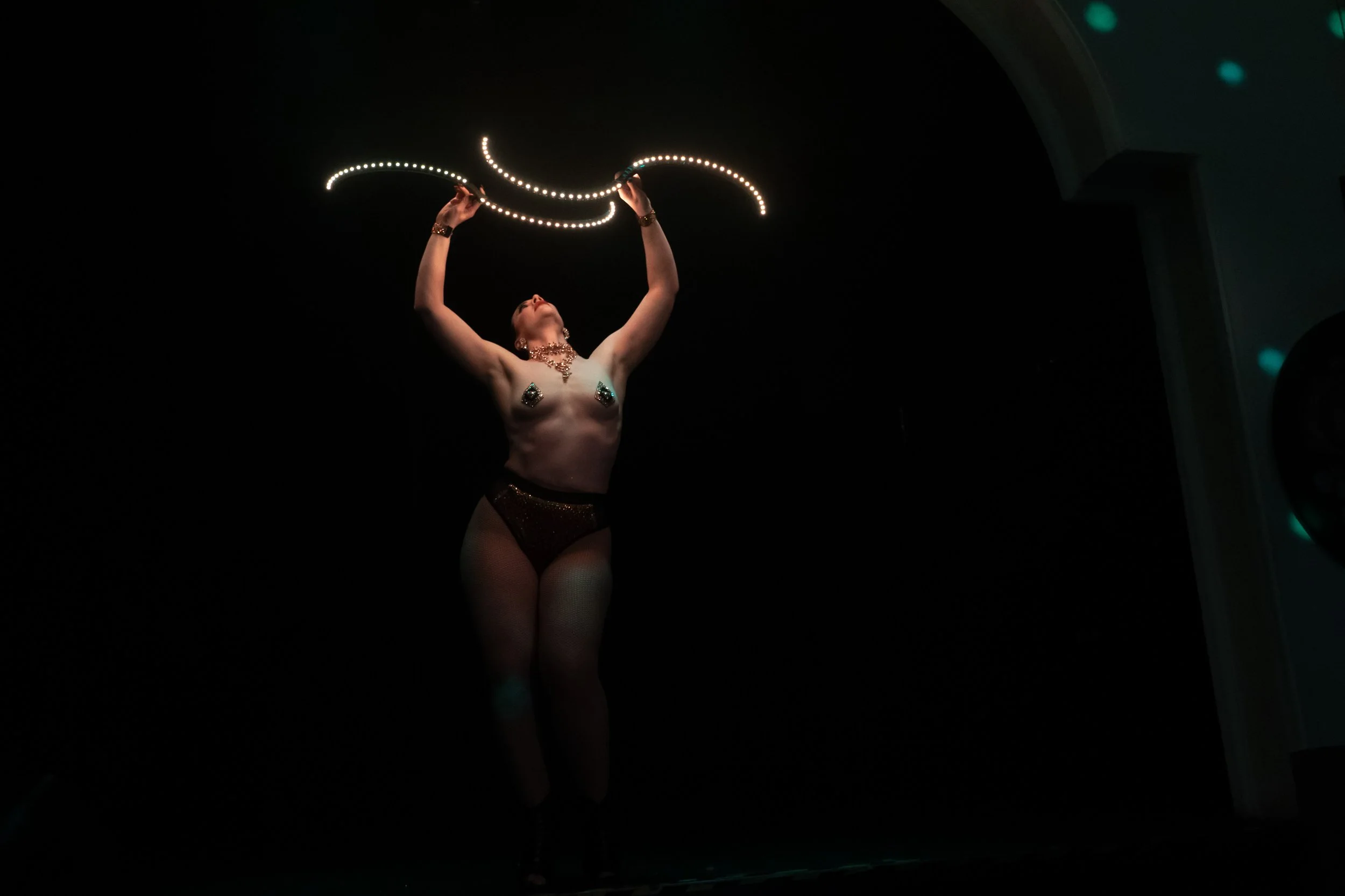 Person performing a light show with LED poi in a dark room, dressed in black underwear and jewelry.