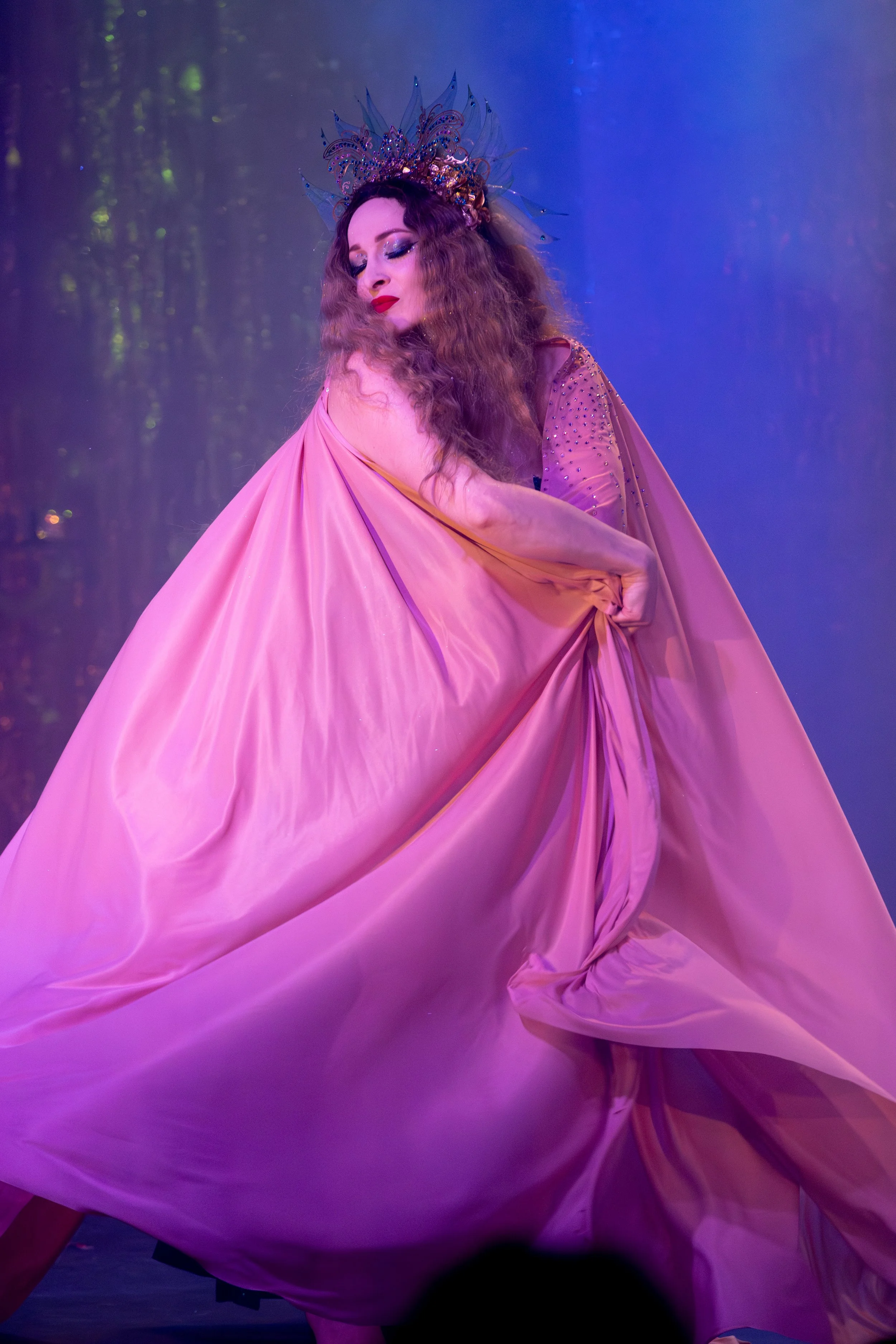 A woman dressed in a flowing pink gown with a crown of dark spikes and beads poses with her eyes closed and head tilted downward.