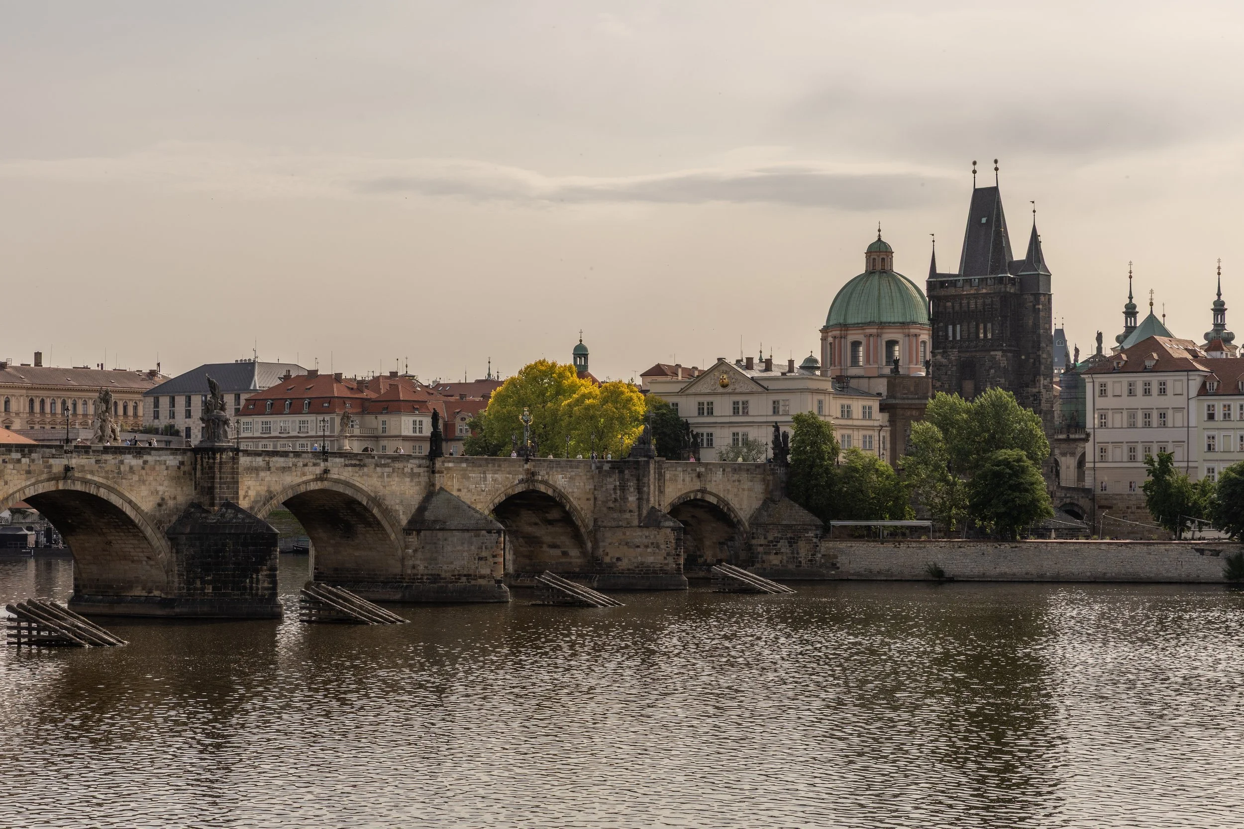 View of a historic European cityscape with a stone bridge over a river, and old buildings including a church with a green dome and a tall dark tower in the background.
