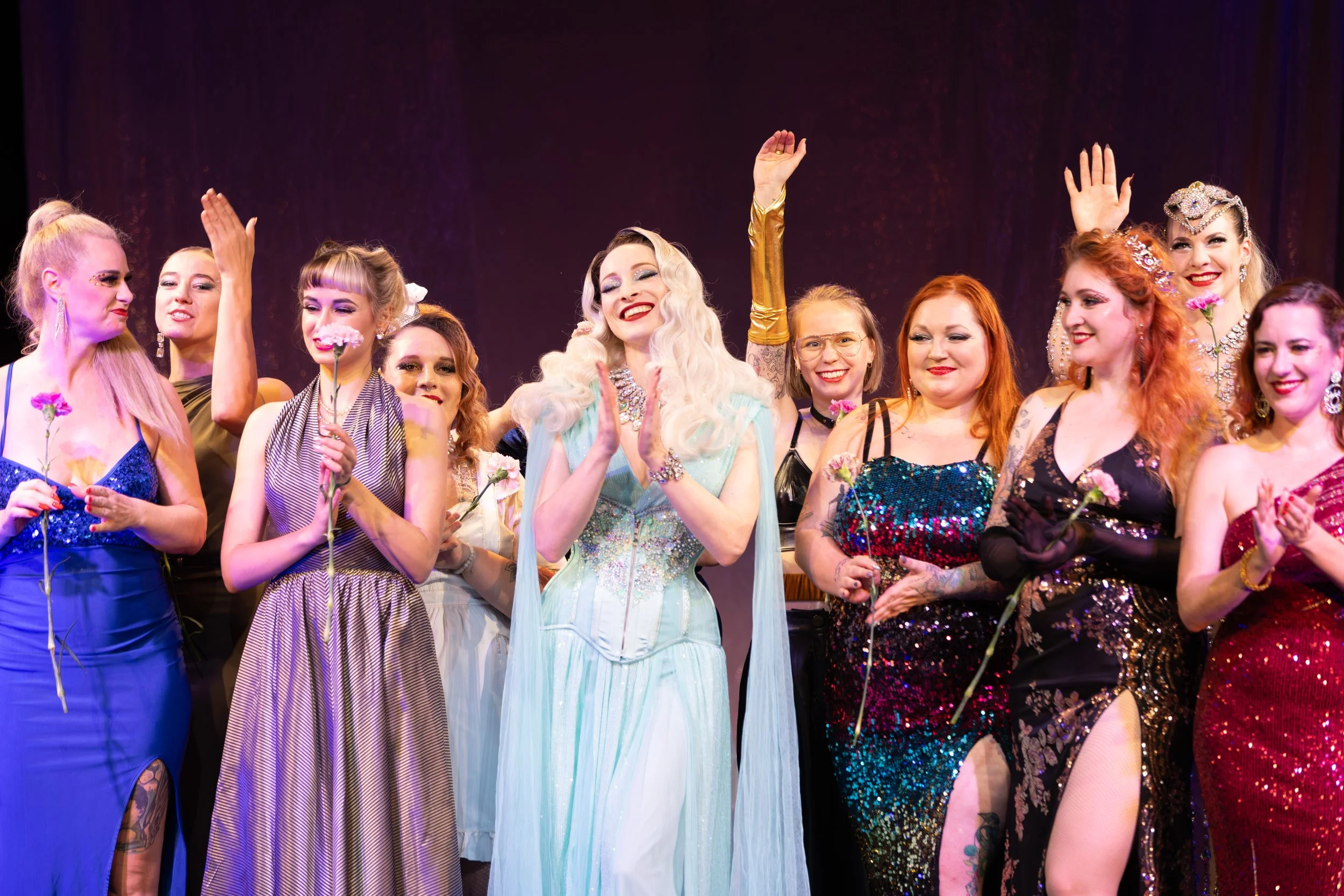A group of women dressed in colorful evening gowns on stage, celebrating with flowers and applause.