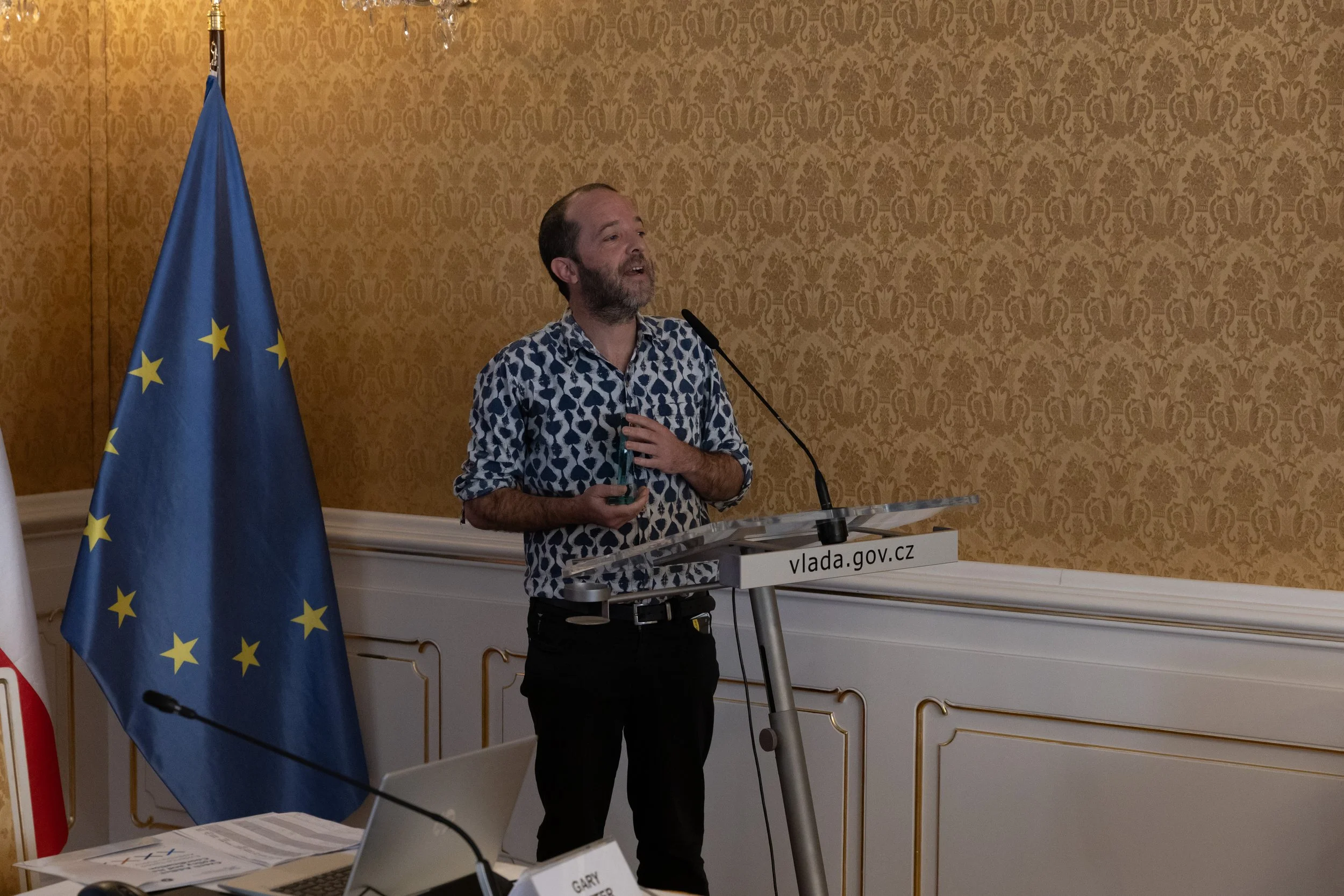 A man with a beard and a patterned shirt is speaking at a podium with a microphone in front of a European Union flag, in a room with ornate wallpaper and white trim.