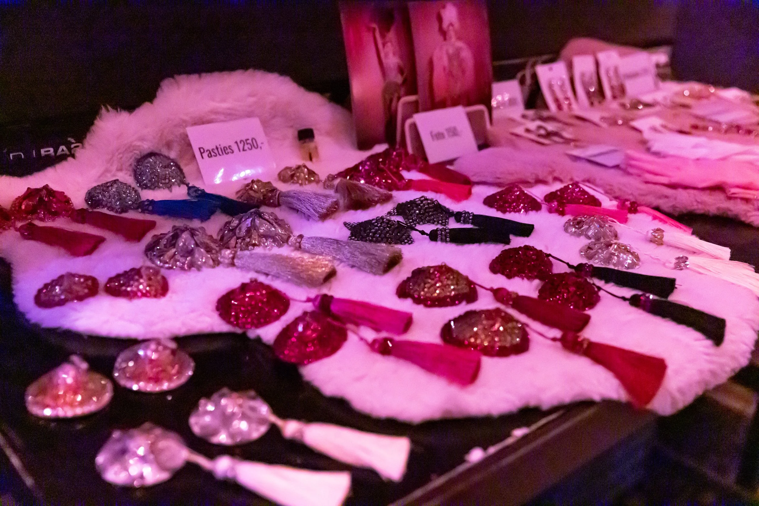 Various earrings and jewelry displayed on a table, with a pink fur cloth and a sign that reads 'Pasties 1250.'