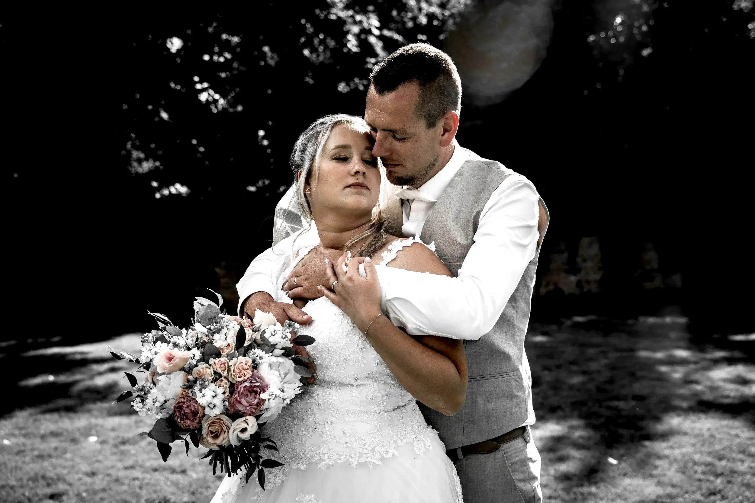 A bride and groom embrace outdoors, with the groom holding the bride and her holding flowers, both with eyes closed in an intimate moment.