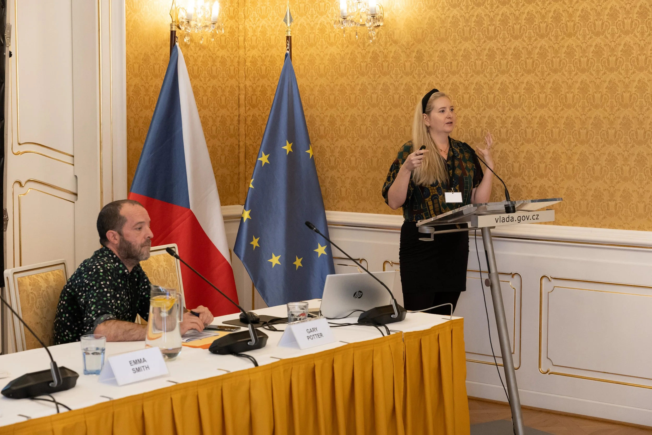A woman is giving a presentation at a podium with the website 'vlada.gov.cz' displayed on it, while two flags, the Czech Republic and the European Union, stand behind her. A man is sitting at a table with a microphone, name tags, water glasses, and d