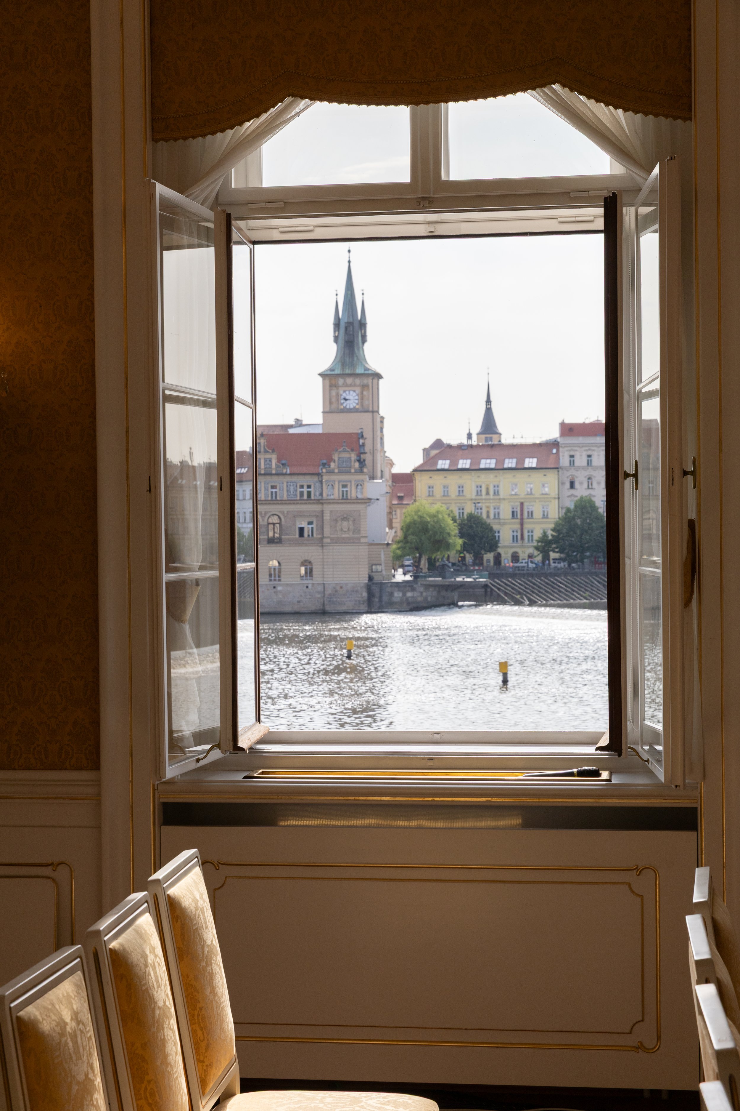 View of historic buildings and a church tower across a river, seen through an open window in an ornate room with gold-trimmed chairs.