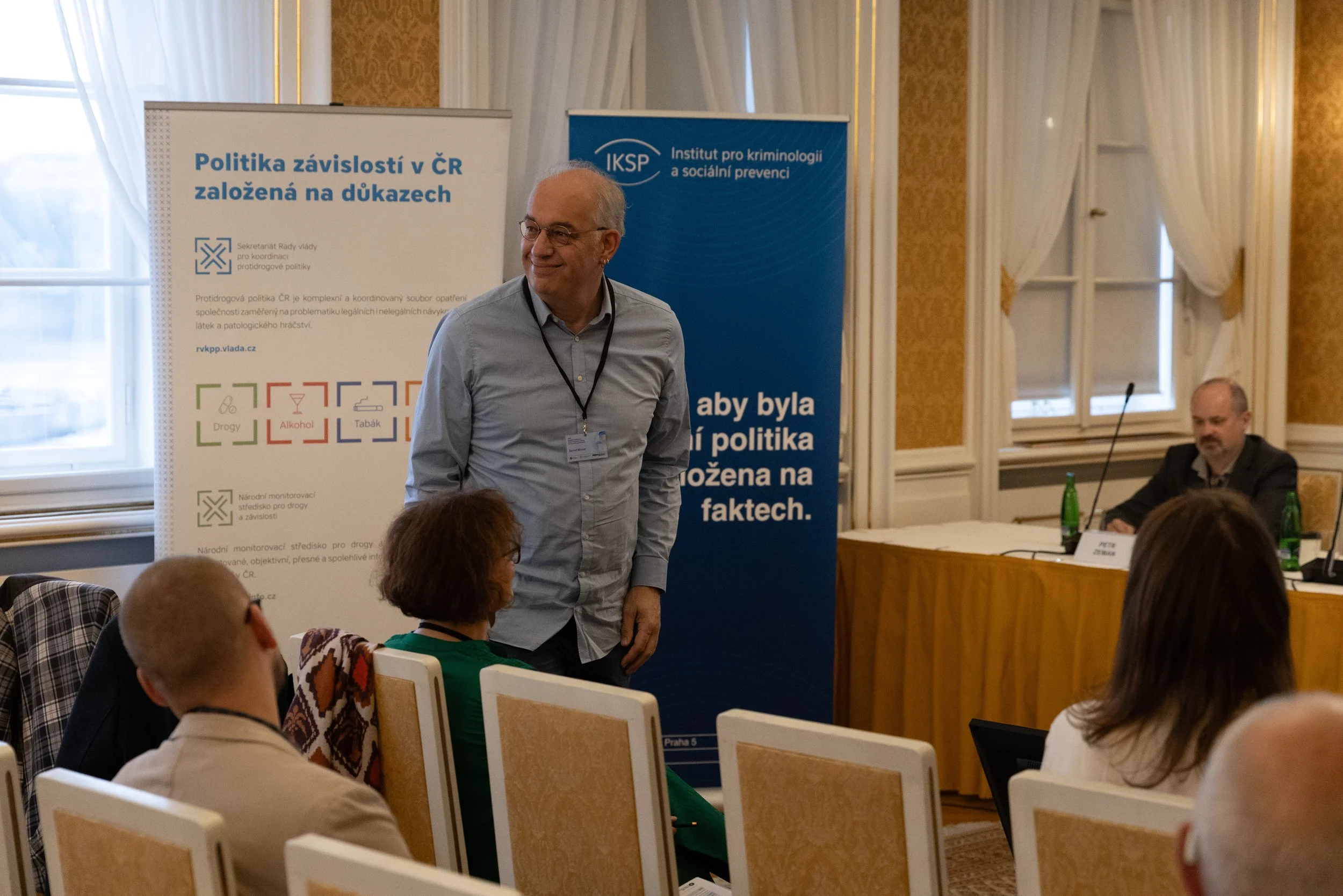 A man stands and speaks at a conference, surrounded by seated attendees. Behind him are banners with text in Czech about addiction policies and social prevention.