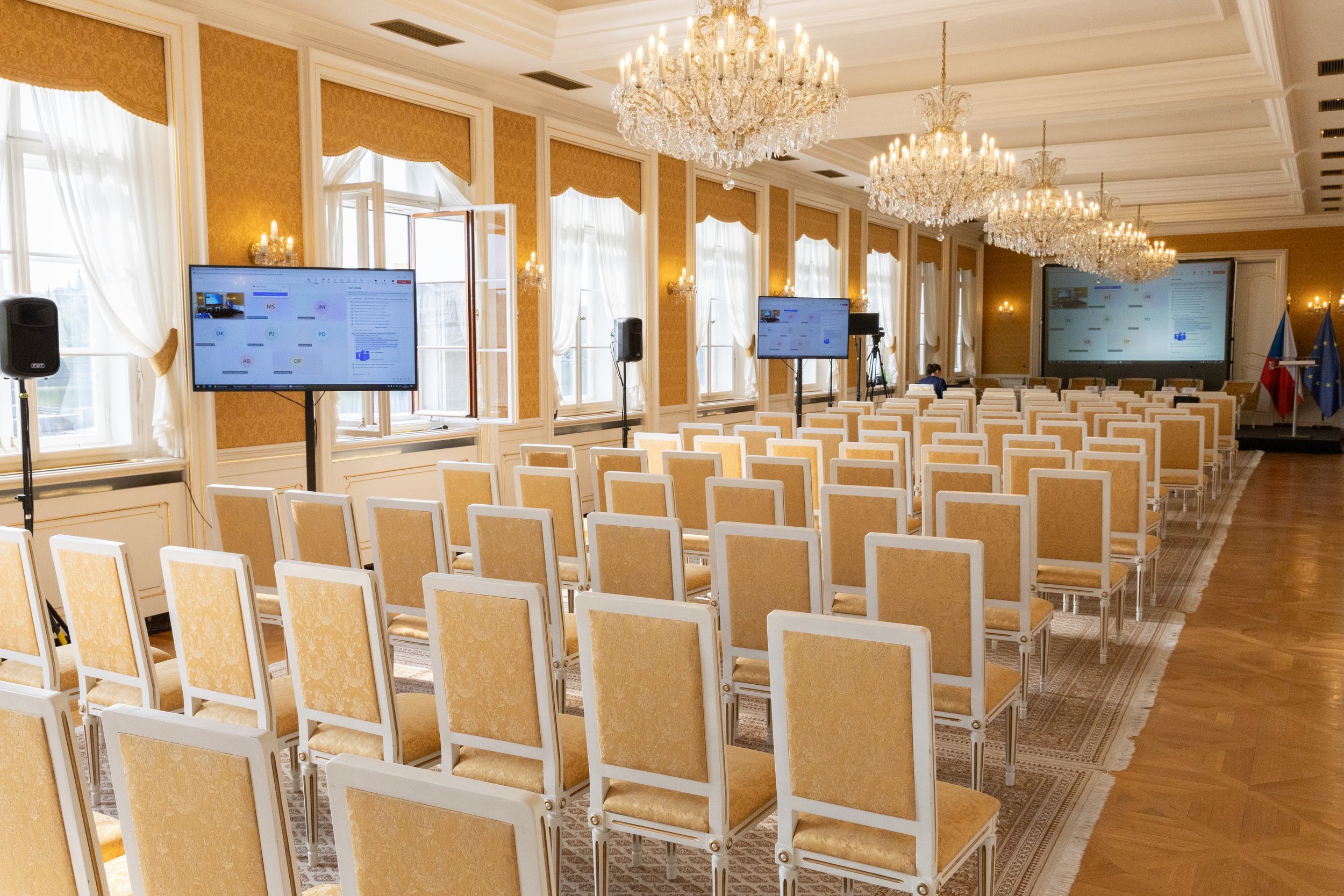 A conference room with rows of yellow and white chairs facing large screens and chandeliers on a decorated ceiling.