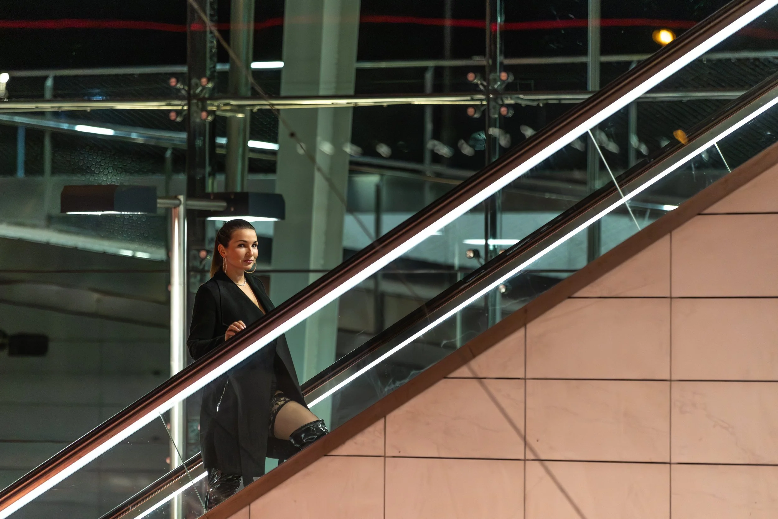 A woman with long hair, dressed in black, sitting on an escalator in a modern building with glass and metal architecture at night.
