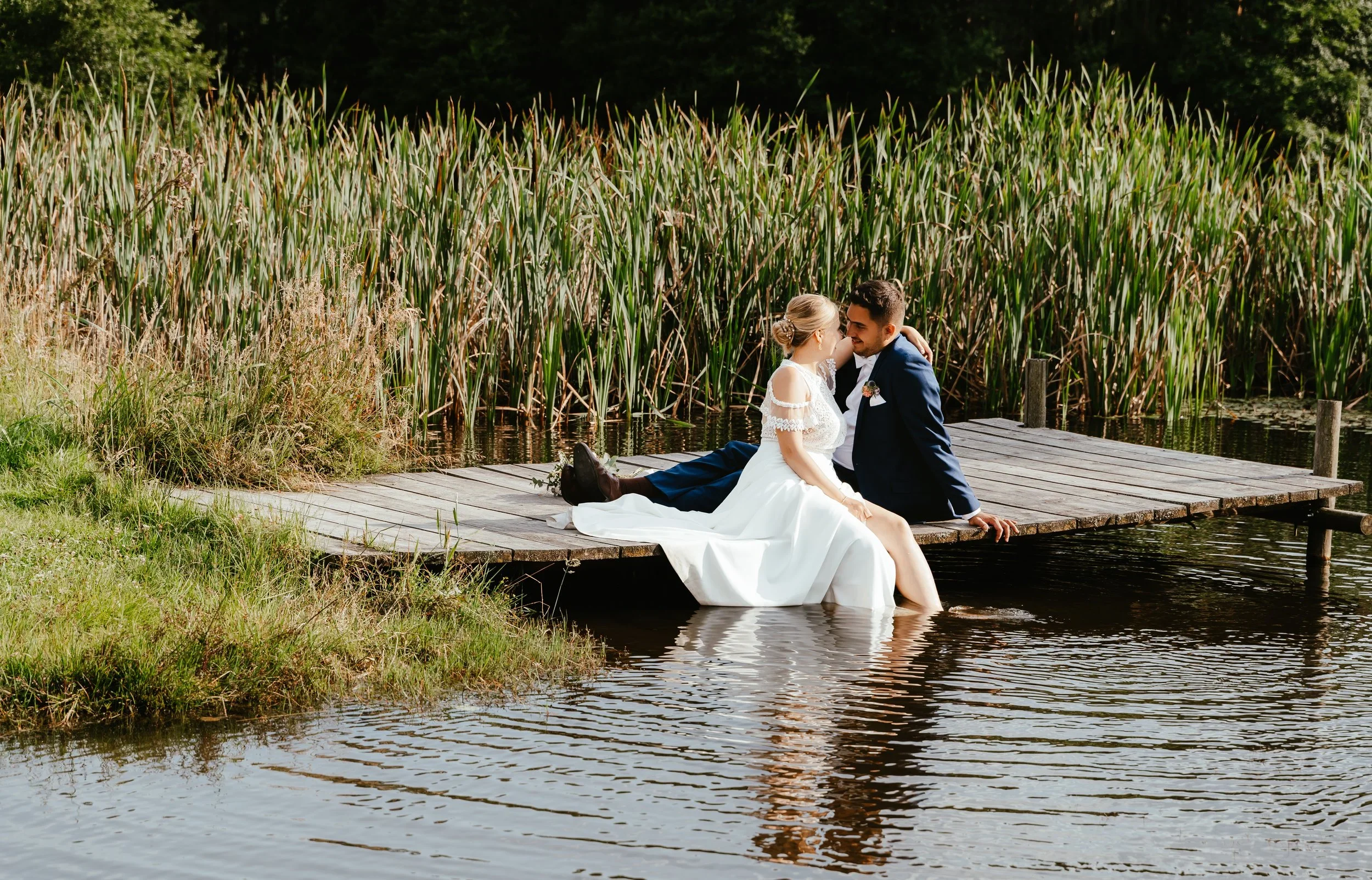 A bride and groom sitting on a wooden dock over a pond, with tall reeds in the background, sharing a romantic moment.