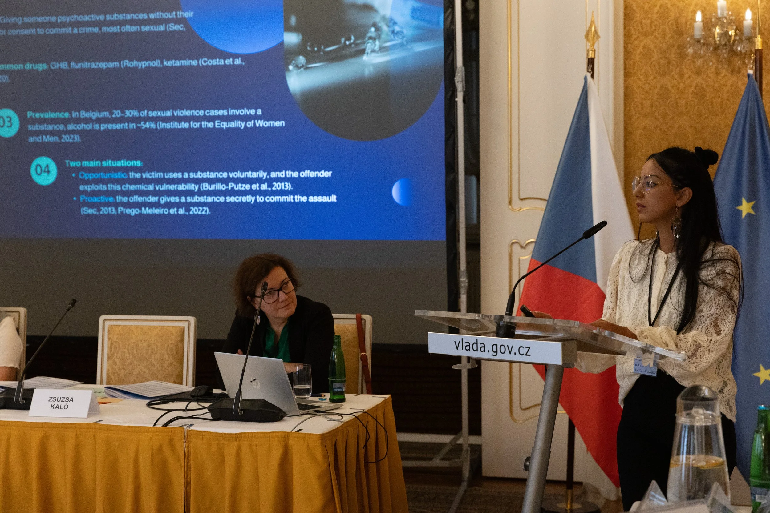 A woman is giving a presentation at a conference, standing behind a podium with a microphone, with the Czech flag and the European Union flag behind her. Two women are seated at a table, one of whom is looking at her, with presentation slides project