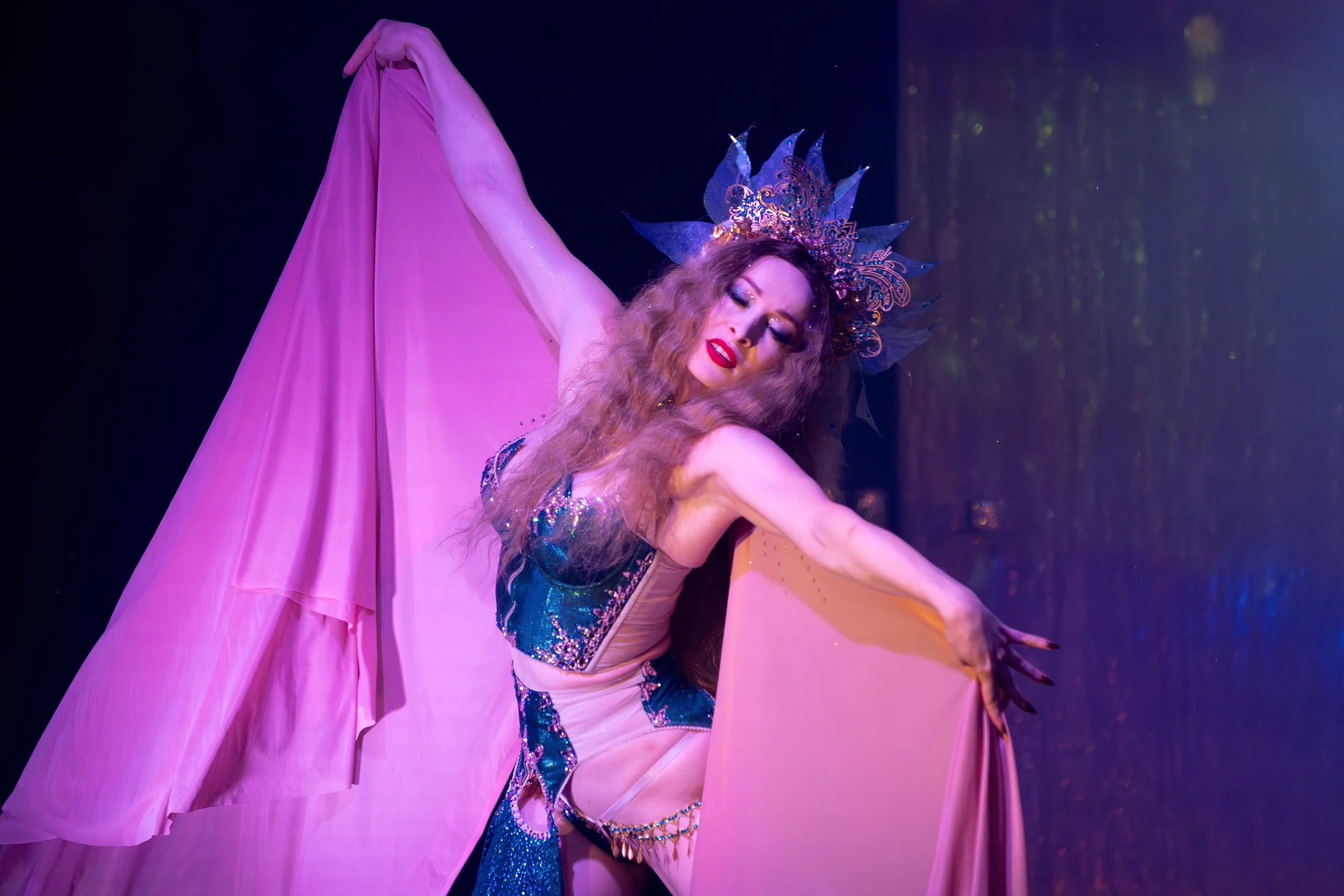 A performer dressed in a vibrant costume with a crown-like headpiece, striking a graceful pose with a pink flowing fabric, on stage with dramatic lighting.