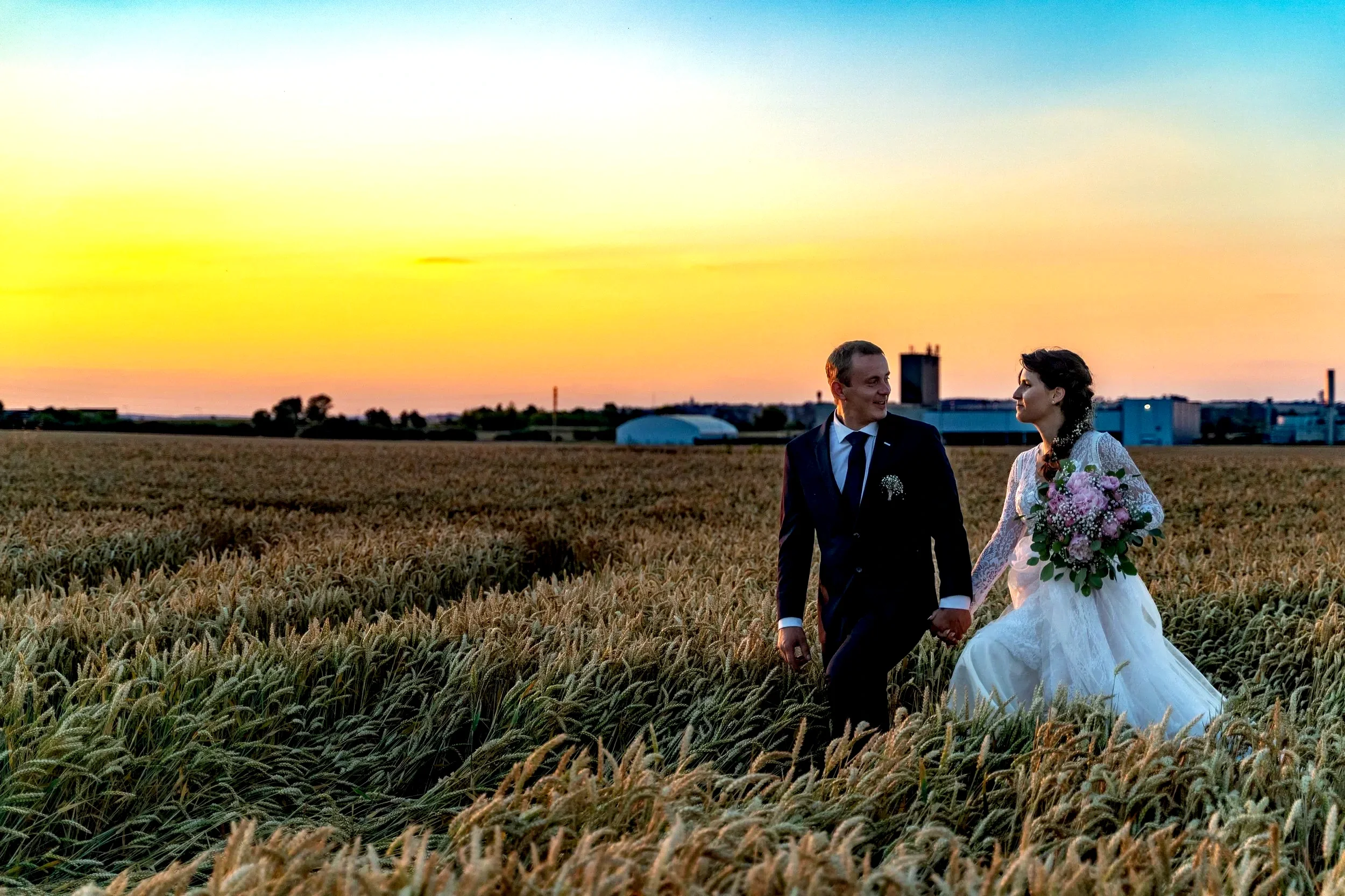 A bride and groom walking hand in hand through a wheat field at sunset, with industrial buildings in the distance.