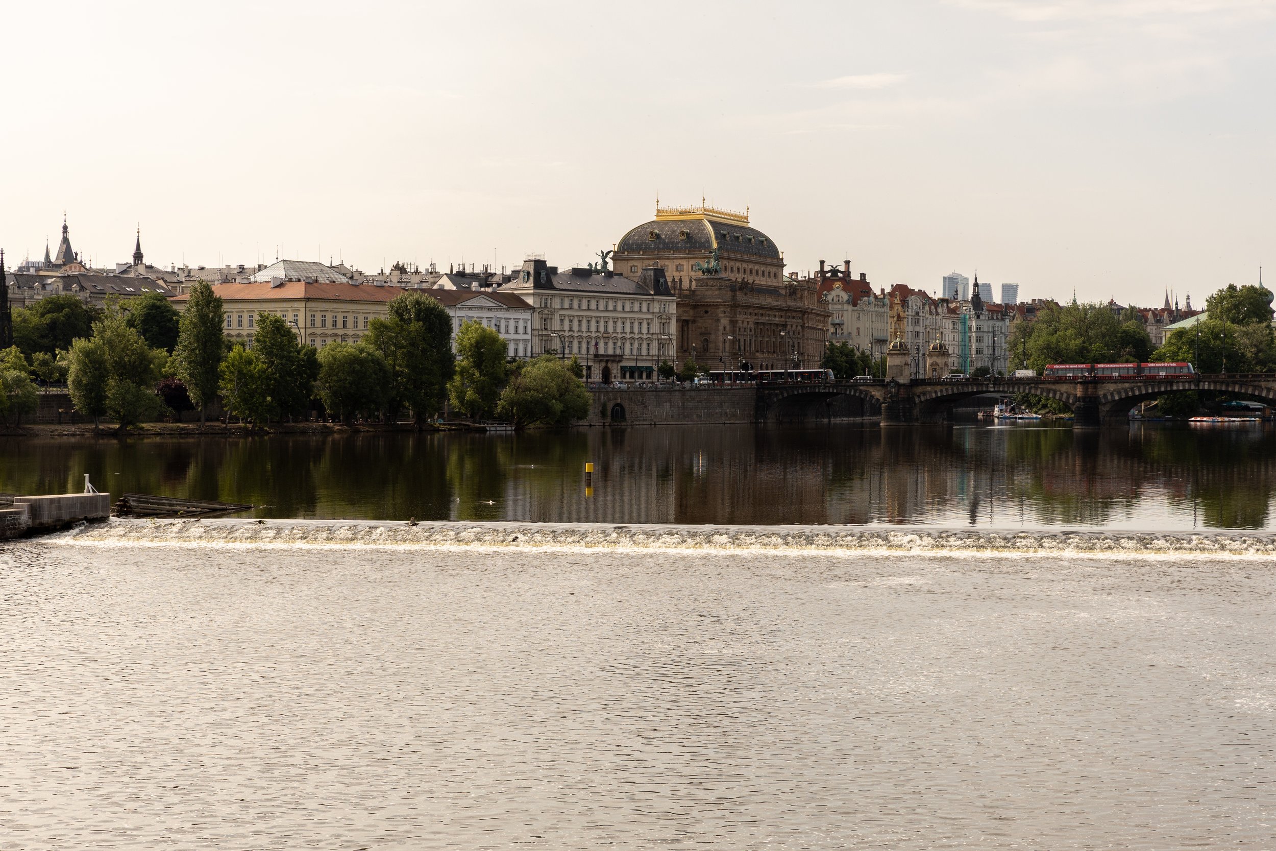 View of a cityscape across a river with historic buildings, trees, and a bridge, under a partly cloudy sky.