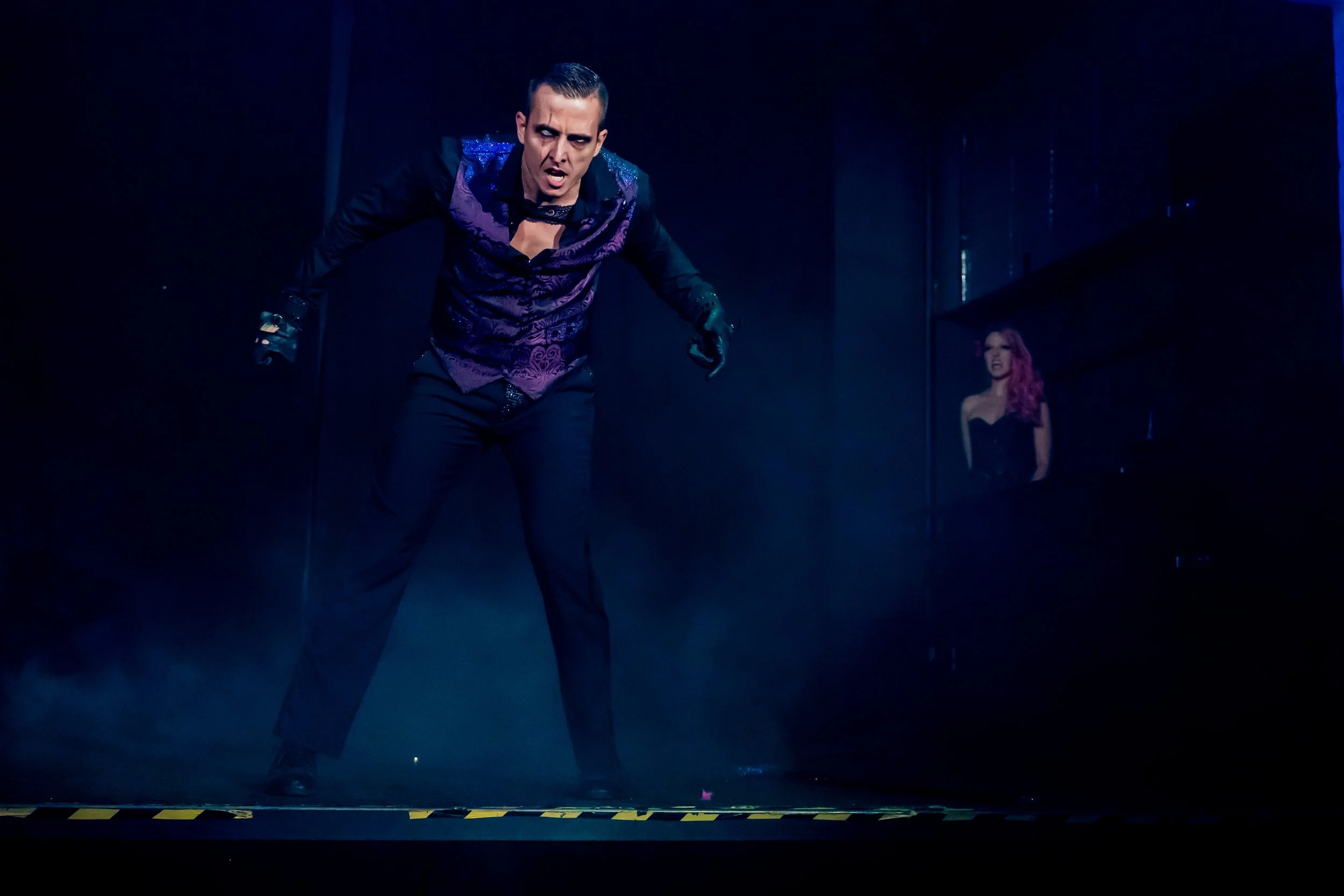 A man in gothic attire, with makeup and dark clothing, appears to be performing on stage with a serious expression. In the background, a woman with pink hair and a black dress is visible, looking at the man.
