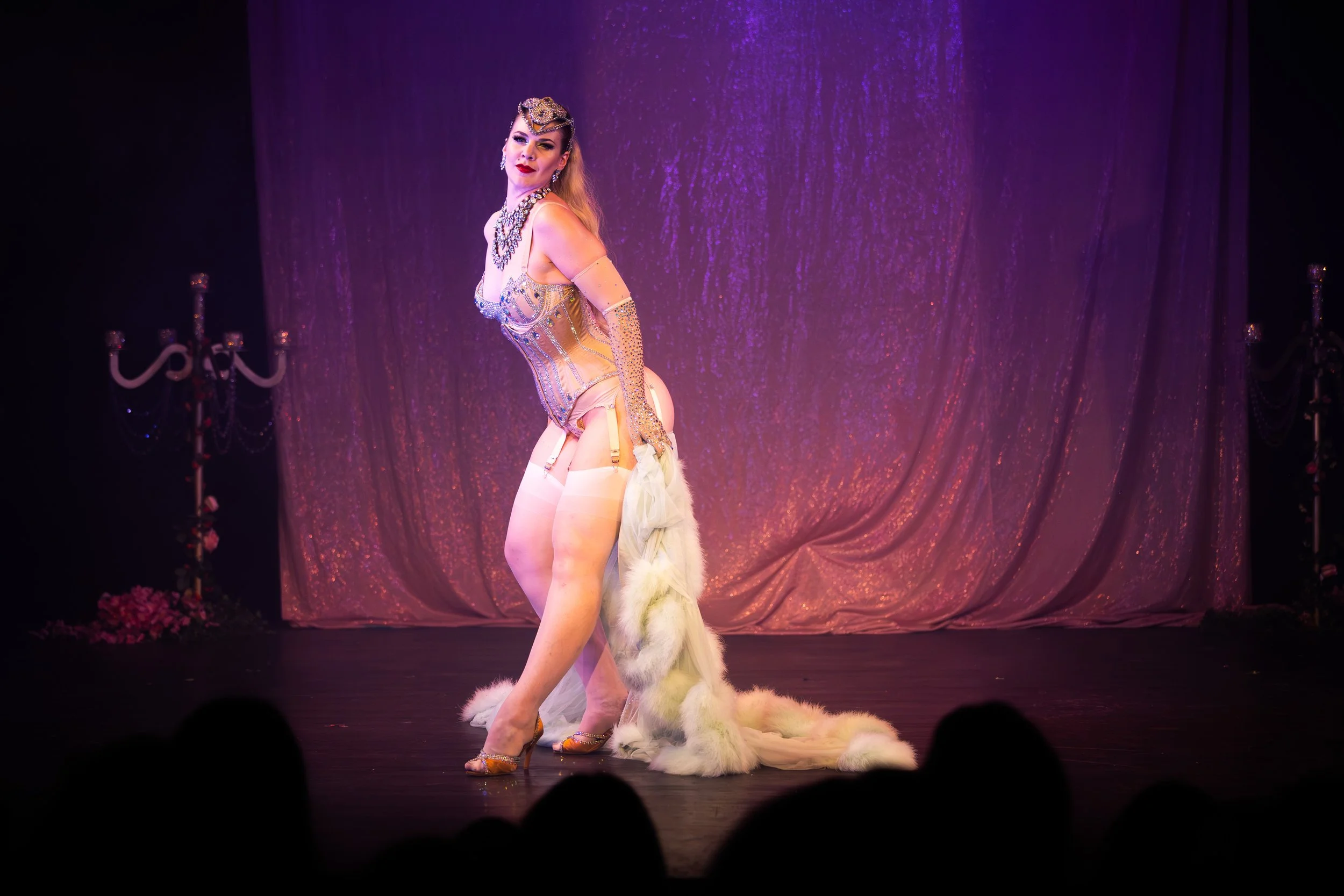 Performer on stage wearing a glamorous costume with a long fur train, posing with confidence against a purple curtain backdrop, with audience silhouette in foreground.