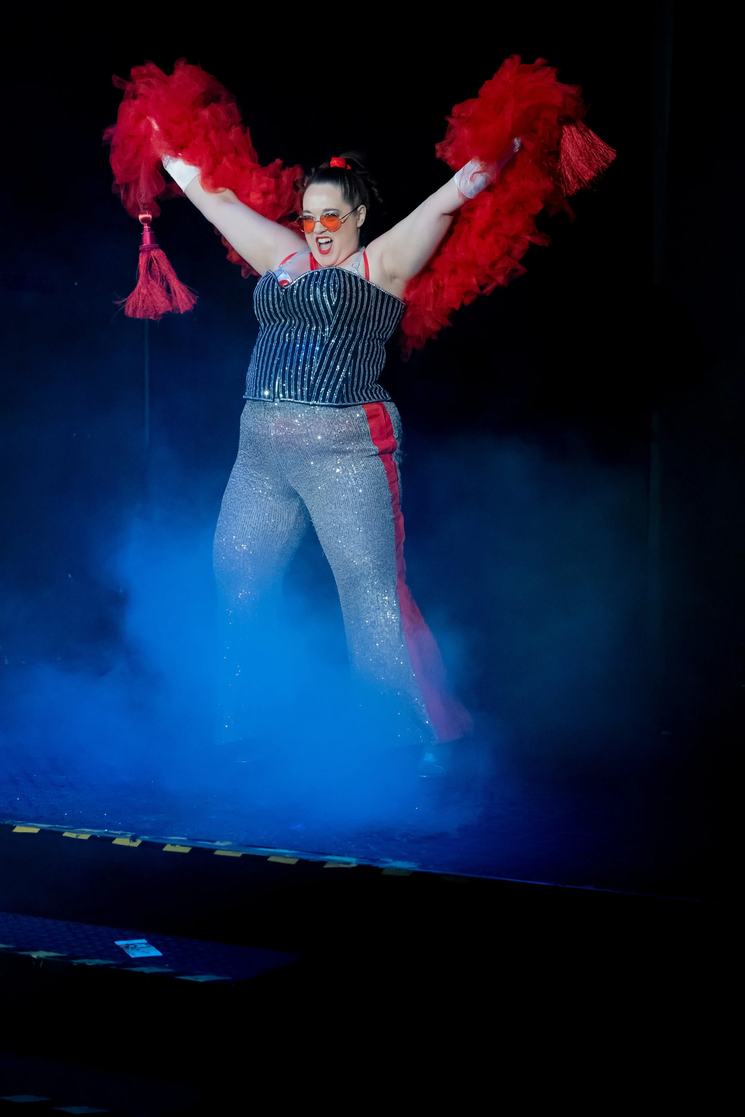Performer on stage wearing glitter pants, a striped top, and red feathered jacket, with red sunglasses, smiling and raising arms.