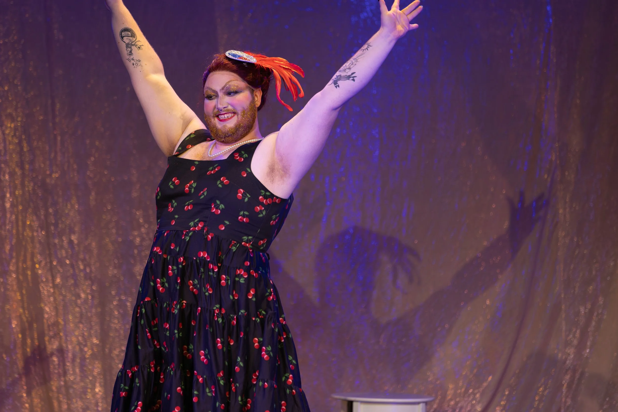 Performer with red hair, beard, and tattoos on arms in a cherry-patterned dress, smiling with arms raised on stage with sparkly backdrop.