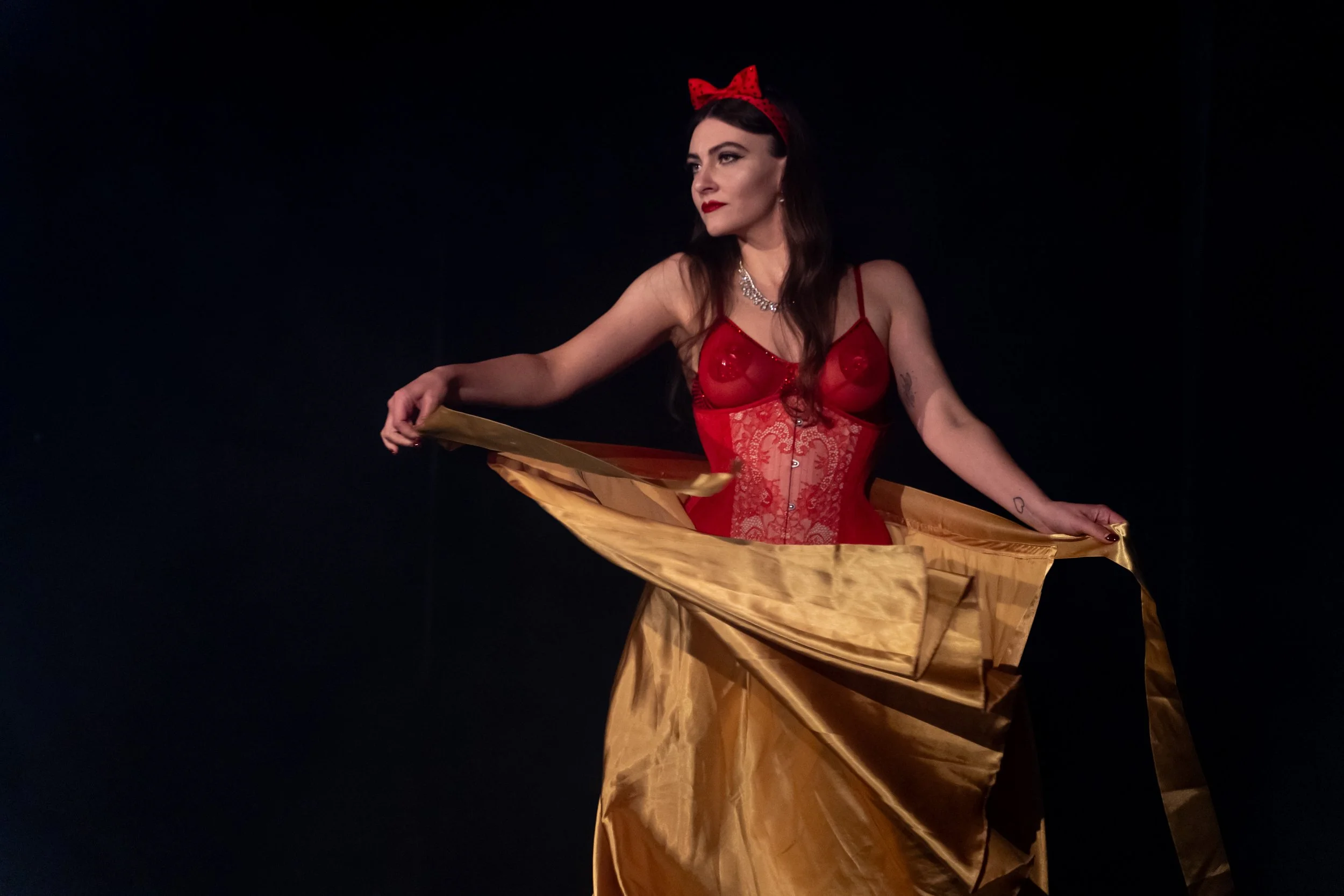 A woman dressed in a red corset top with lace details, wearing a red bow headband, and holding a gold fabric or skirt against a black background.