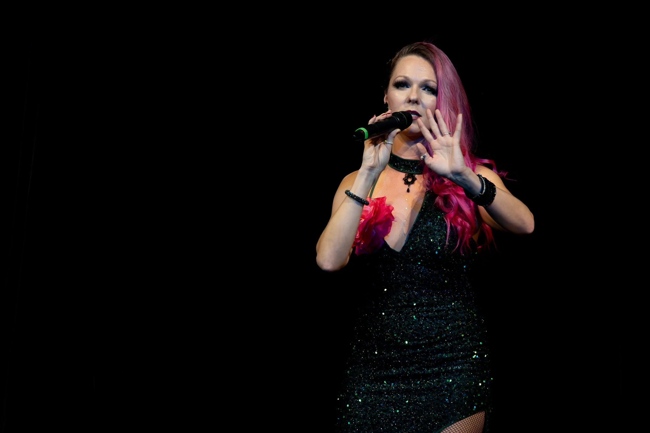 A woman with pink hair performing on stage, holding a microphone and wearing a black glittery dress with a floral detail on the shoulder, against a black background.
